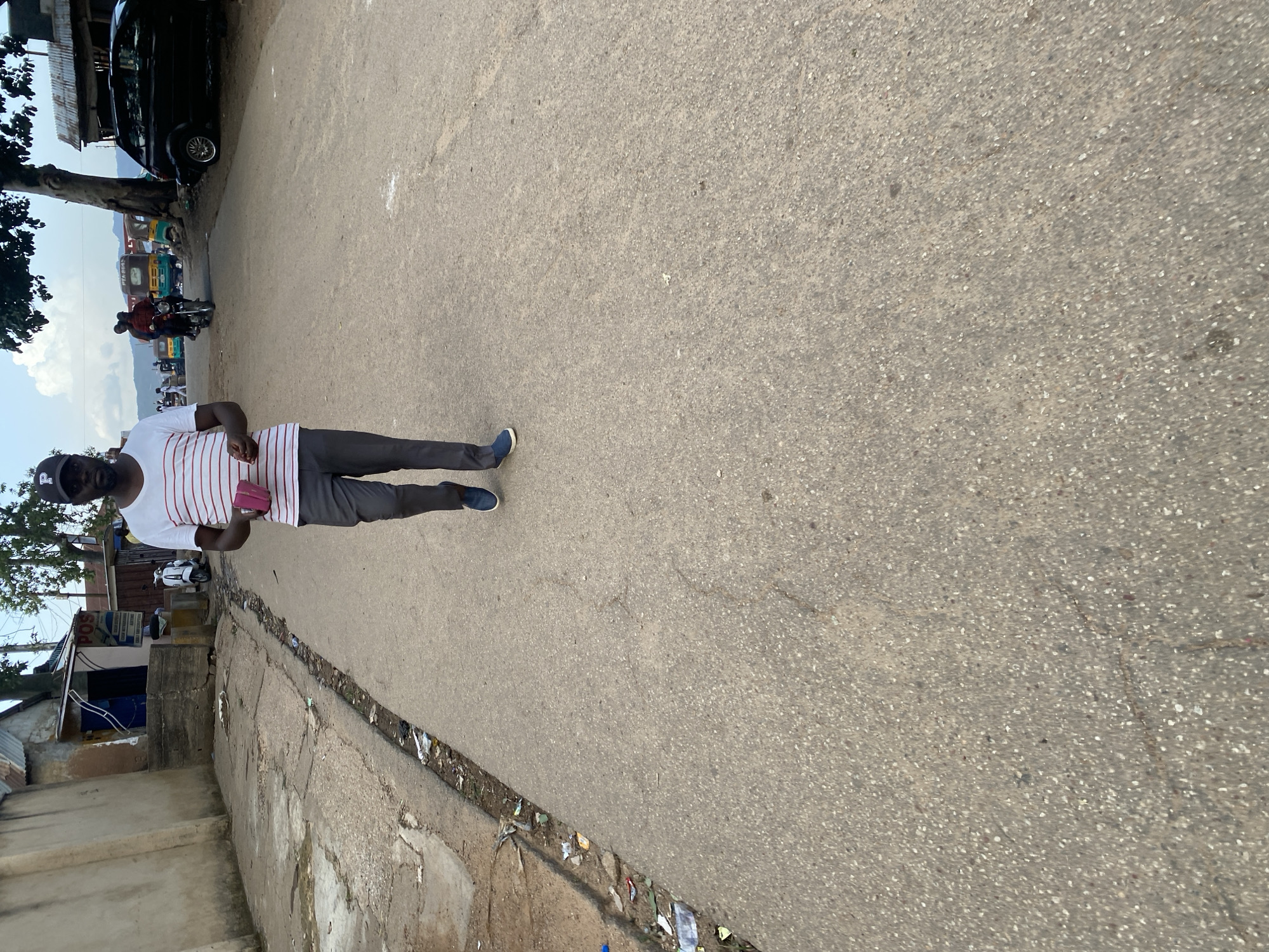 A man wearing a white and red striped t-shirt and gray pants walks down a paved street. He is wearing a dark baseball cap with a white "P" logo. He holds a small pink object in his right hand. The street is lined with buildings on the left and trees and vehicles on the right. In the background, several motorbikes and a black car are visible. The environment appears to be an urban setting.
