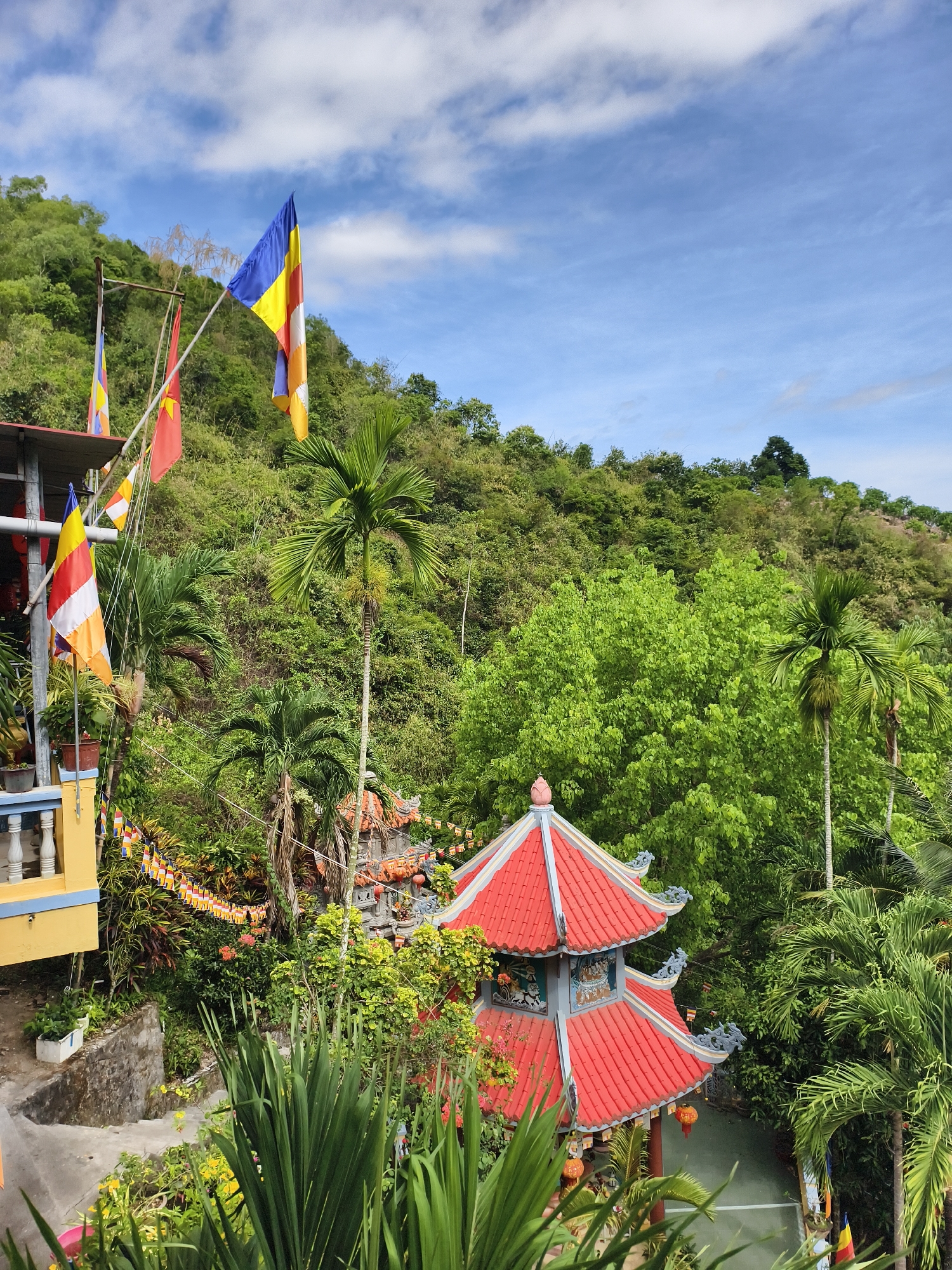The image shows a vibrant scene in what is possibly the city of Tay Nha Trang Ward, Vietnam. In the foreground, a red-roofed pagoda is partially obscured by lush greenery. To the left, a building with a yellow and blue balcony is visible. Several Buddhist flags in bright yellow, red, blue, white, and orange are hoisted on poles, fluttering against the backdrop of a partly cloudy, blue sky. The background is dominated by a hill densely covered in green trees, suggesting a tropical location. The environment suggests a serene and possibly religious setting.