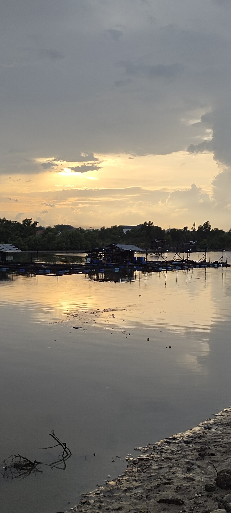 This image depicts a serene sunset scene at a floating fish farm located in the city of Lhokseumawe, Indonesia. The sky is filled with dramatic, grey clouds, but a patch of warm, golden sunlight breaks through, illuminating the water with a soft glow. In the foreground, the calm water reflects the sky and the structures on the water. A muddy shoreline with scattered debris is visible on the right. In the middle ground, a series of interconnected floating rafts, likely part of the fish farm, are dotted with small, weathered structures, some appearing to be sheds or living quarters. The rafts are supported by blue barrels, and wooden posts rise from them, possibly for nets or equipment. A line of dense green trees forms the background, separating the water from the distant, hazy landscape where a few buildings are discernible. The overall atmosphere is peaceful and tranquil, characteristic of a rural or coastal Indonesian setting at dusk.