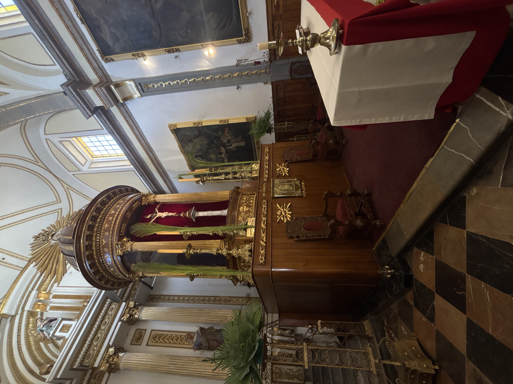 This indoor, medium shot depicts the interior of a church, likely located in Puebla, Mexico, based on its architectural style. The image is taken from a low angle, focusing on a large wooden structure that serves as an altar or display for religious artifacts. This structure is adorned with gold accents and carvings, and features a central crucifix with palm fronds.
In the background, a red and white altar cloth is visible, along with a framed painting on the wall. The church interior features ornate arches, gilded columns, and religious statues. The floor is tiled in a black and brown checkered pattern. The lighting is soft and diffused, highlighting the gold details and creating a reverent atmosphere. There are no people visible in the frame. The architectural details and religious artifacts suggest a Catholic church with a rich history and tradition.