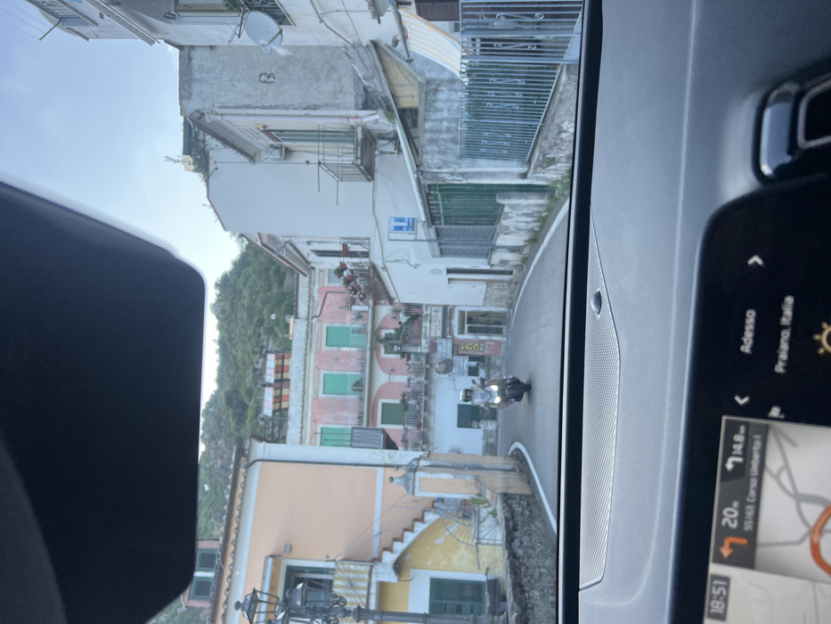 The image provides a direct forward view from inside a vehicle onto a street in Cetara, Italy, at 18:51, according to the visible navigation screen which also indicates "53163 Corso Umberto I, Cetara, Italia" and a destination of "Praiano, Italia."
The foreground displays the vehicle's dark dashboard and an illuminated digital screen showing navigational information, time, and location data.
The central scene depicts a paved, narrow road curving slightly to the left. An individual wearing a light-colored helmet and clothing is operating a light-colored scooter or motorcycle, traveling towards the viewer along the road.
Buildings line both sides of the street. On the left, a light peach-colored building features a prominent exterior staircase with yellow steps and white railings. An ornate, dark-colored street lamp is positioned in front of this structure. Further back and slightly elevated, a pink building is visible, characterized by multiple windows with green shutters and balconies adorned with red flowers.
On the right, structures are primarily off-white, with some featuring balconies and dark railings. Green metal fencing secures the lower portion of a building. A blue and white sign, likely indicating services, is mounted on a wall. A striped, yellow and white awning is partially visible on the far right. A satellite dish is mounted on an upper building facade on the right side.
The background is dominated by steep, green, tree-covered hills rising behind the urban structures. A large, multi-colored striped structure is visible on a distant rooftop on the hillside. The overall light suggests late afternoon or early evening conditions.