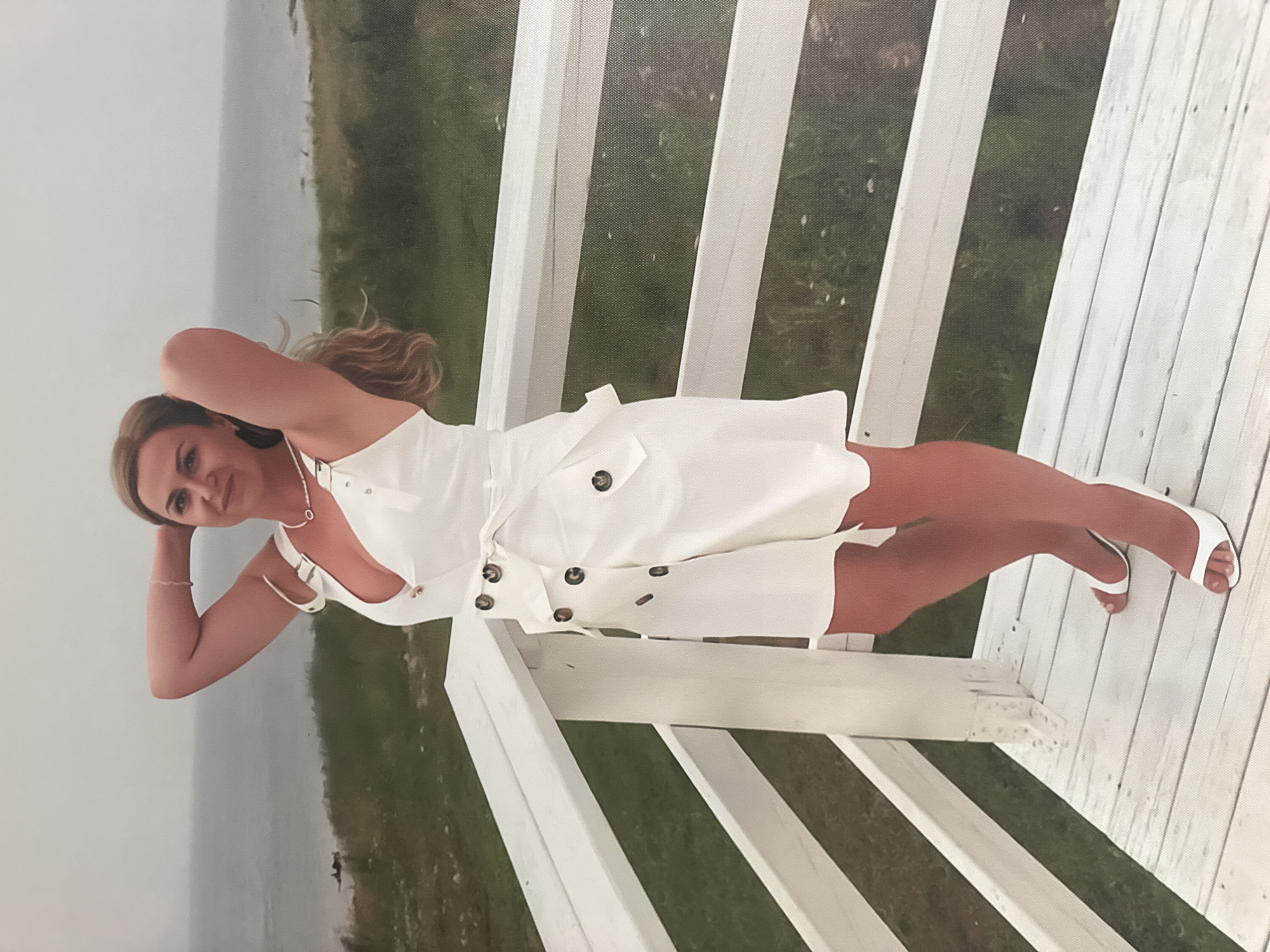 A full-body, slightly low-angle shot shows a Caucasian woman standing on a white wooden balcony railing. She is wearing a white sleeveless dress with a plunging neckline and a belted waist. Her hands are raised to her head, and her hair is blonde and wavy. She is wearing white high-heeled shoes. The background consists of a body of water, green grass, and a cloudy sky. The balcony railing is made of white painted wood and has several horizontal slats. The image appears to have been taken outdoors during the day. There is no indication that this image is located in Bratsk, Russia.