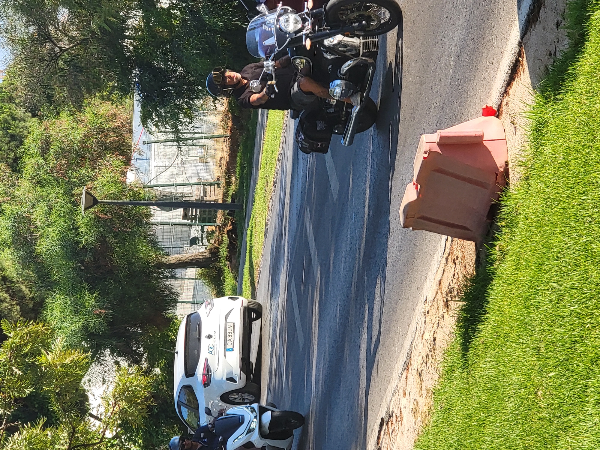 In Quarteira, Portugal, a man rides a black motorcycle down a paved road lined with green grass and trees. He wears a black t-shirt, grey shorts, a helmet, and goggles. The motorcycle has a large windshield and saddlebags. A white car with the license plate "CA 99 JV" is parked on the side of the road, and a white scooter is parked behind it. A red traffic barrier is also visible on the grass. The sun is shining, suggesting it is daytime and the weather is clear. The scene appears to be a sunny day in a suburban or park-like area.