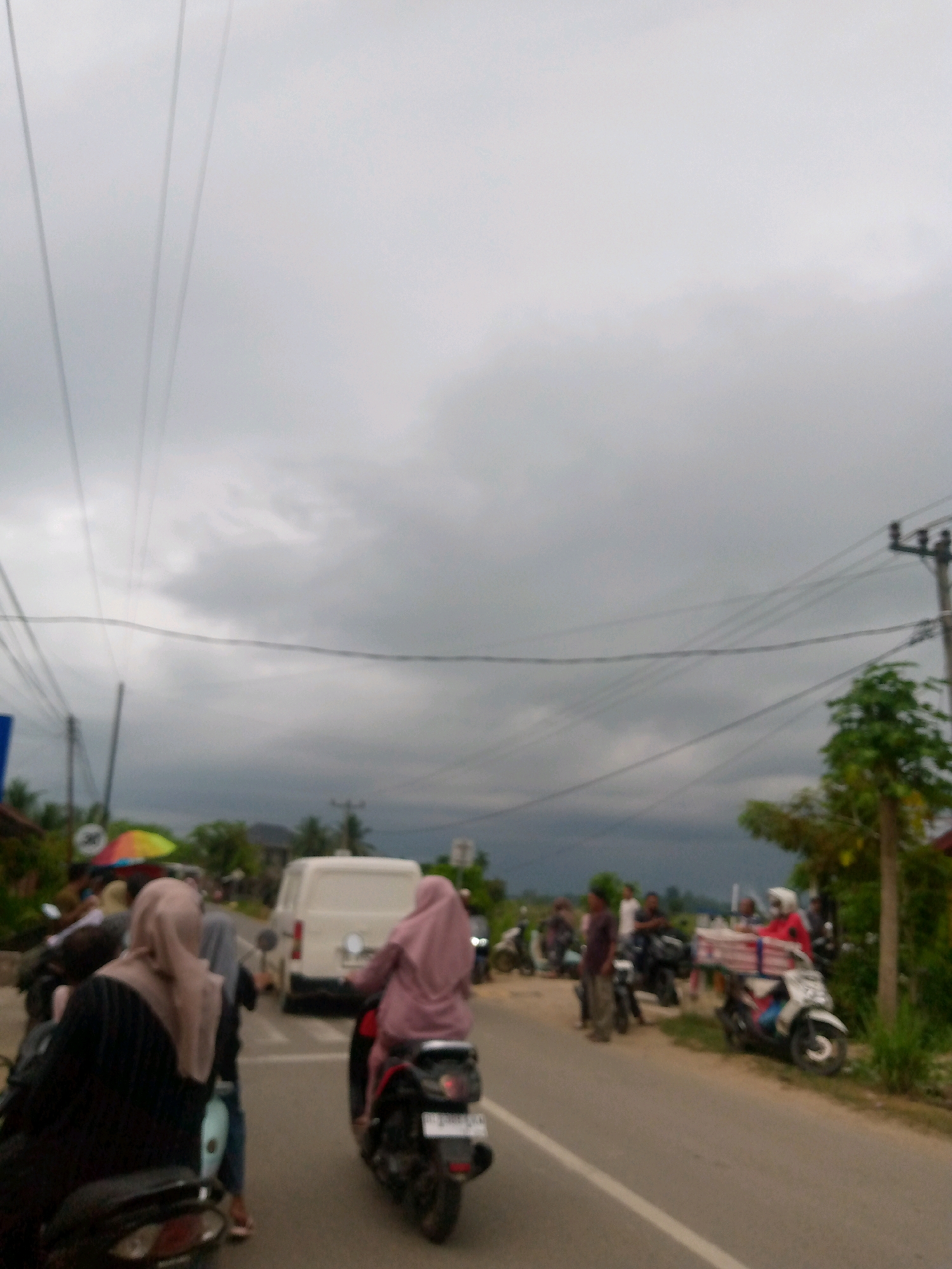 The image depicts a street scene in what appears to be a rural or suburban area, possibly in the city of Mesjid, Indonesia, under an overcast sky. The foreground shows several people on motorcycles, some of whom are wearing headscarves. One person in the immediate foreground is wearing a black top and a light-colored headscarf, seated on a motorcycle. To their right, another person wearing a pink headscarf is riding a motorcycle. Further down the road, a white van is visible, and other motorcycles and people are scattered throughout the scene. Power lines crisscross the sky, and vegetation, including trees and grass, lines the side of the road. The overall impression is of a casual, everyday street with moderate activity.