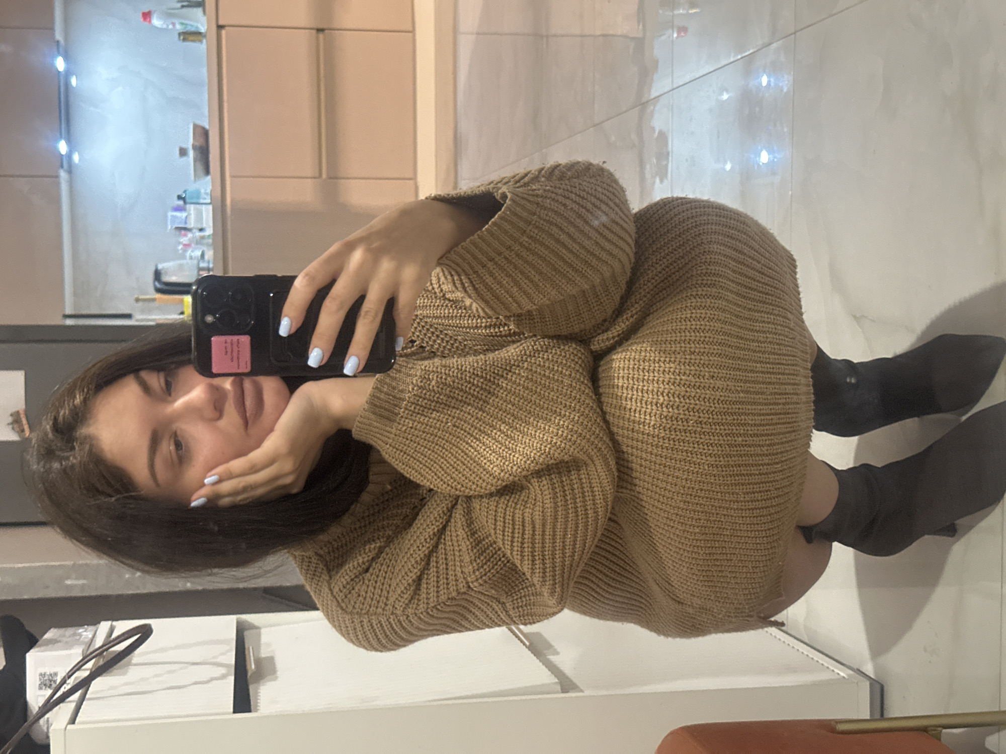A woman is taking a selfie while kneeling indoors. She is wearing a large, tan-colored sweater dress and dark boots. The background includes cabinets and what appears to be tiled flooring with reflective lights. A white shelf is visible in the foreground. No specific identifying features confirm the location as Khimki, Russia.
