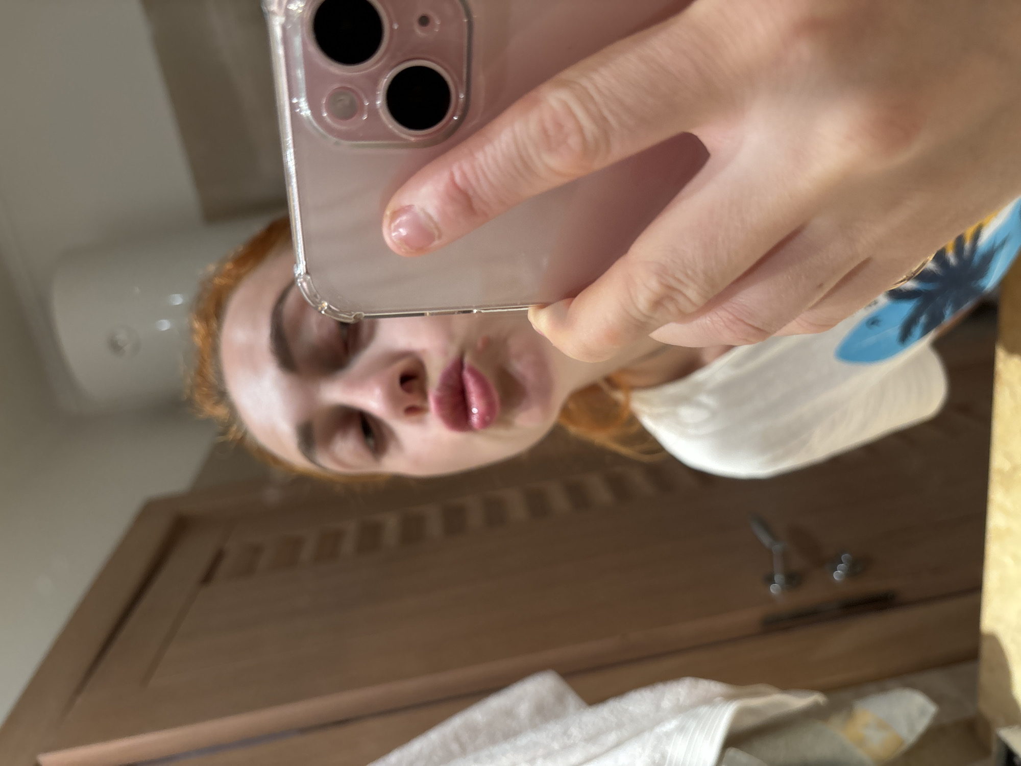 A woman with red hair is holding a phone and taking a selfie in what appears to be a bathroom. The phone has a clear case. The woman is wearing a white t-shirt with a blue design. The bathroom has a wooden door, white towel, and a white bathroom fixture on the wall. The location in Budva, Montenegro is unknown.