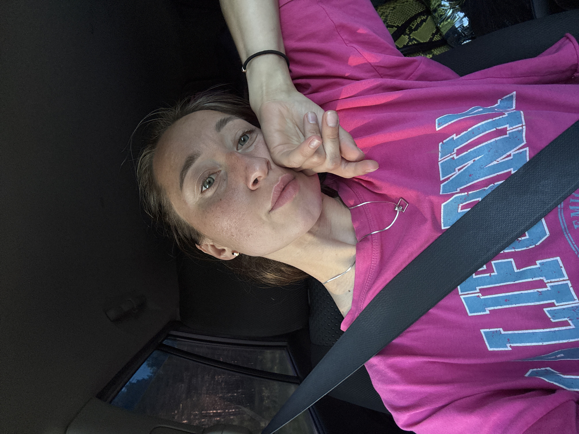 A young adult female is depicted in a close-up, frontal selfie taken from the front passenger seat of a vehicle in Шумиха, Russia. She is wearing a bright pink long-sleeved t-shirt featuring distressed light blue lettering that spells "CALIFORNIA." A black seatbelt is positioned diagonally across her chest and lap, securing her. Her left hand supports her head, with her elbow resting out of frame to her right. The subject has light skin, visible freckles across her nose and cheeks, and light-colored eyes. She wears a silver chain necklace with a small geometric pendant, a small dark earring in her right ear, and a black elastic hair tie on her right wrist. The vehicle's interior is dark, with a dark headliner and seat upholstery. Through the window to her left, a dark forested landscape is discernible under a dim blue sky, indicating evening or night conditions. In the bottom right corner, a dark green object with a textured, possibly reptilian, pattern is partially visible. The scene is lit softly, highlighting the subject's face and the front of her shirt.