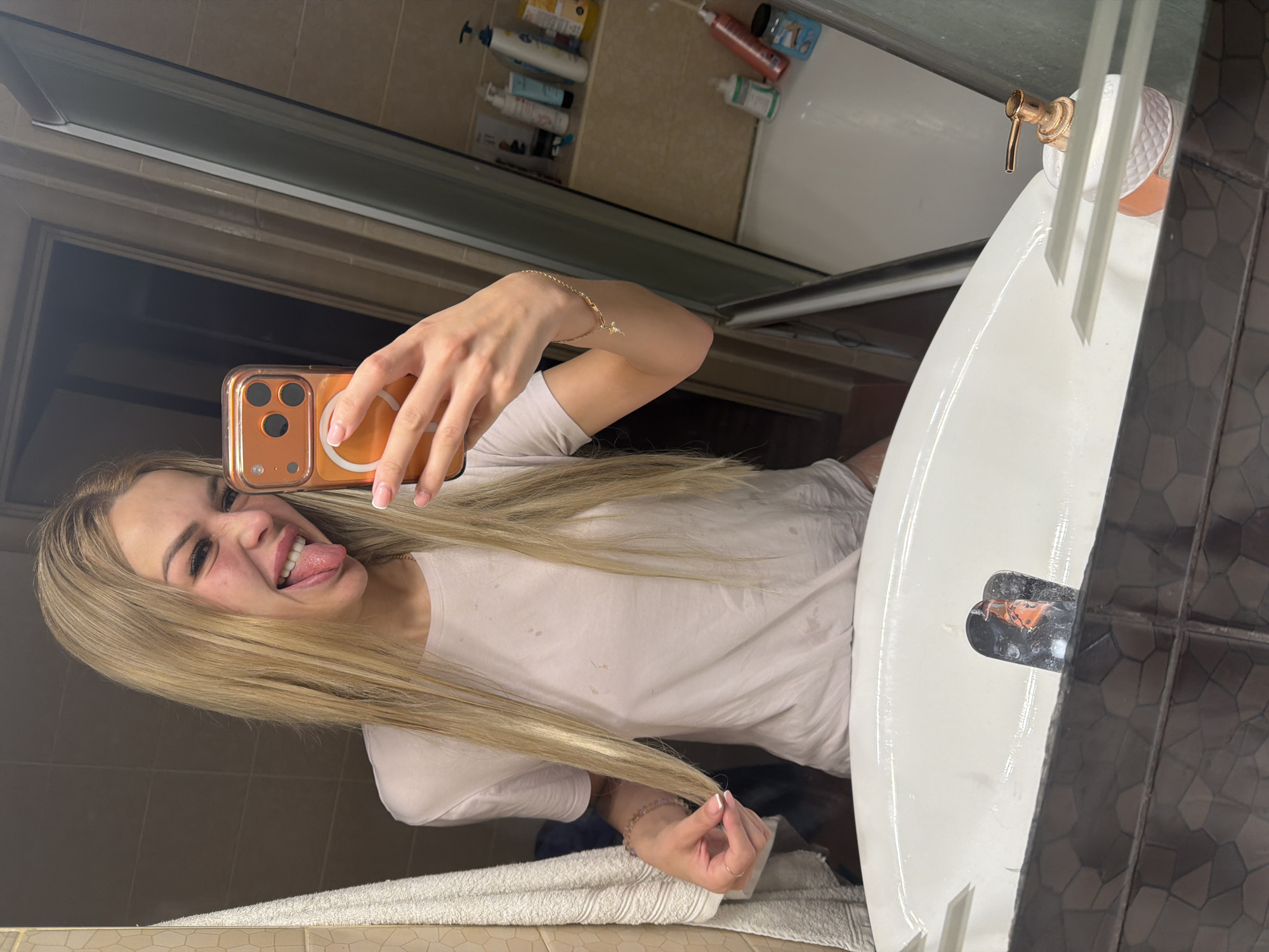 A woman with blonde hair is taking a selfie in a bathroom. She is sticking her tongue out and making a funny face. She is wearing a light pink t-shirt. In the background, there are various toiletries on a shelf and a bathtub. The scene appears to be in a residential bathroom, with tiled walls and a sink in the foreground. There is no indication of the specific location within Kyiv, Ukraine.