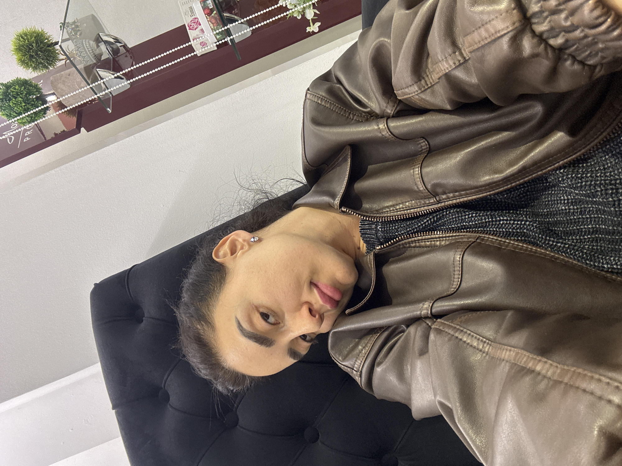 A woman rests on a black tufted headboard, wearing a brown leather jacket over a gray knit top. A shelf above her contains small potted plants and decorative items. The location appears to be indoors, potentially in Tbilisi, Georgia, based on the general aesthetic, but there is no definitive evidence in the image to confirm this.