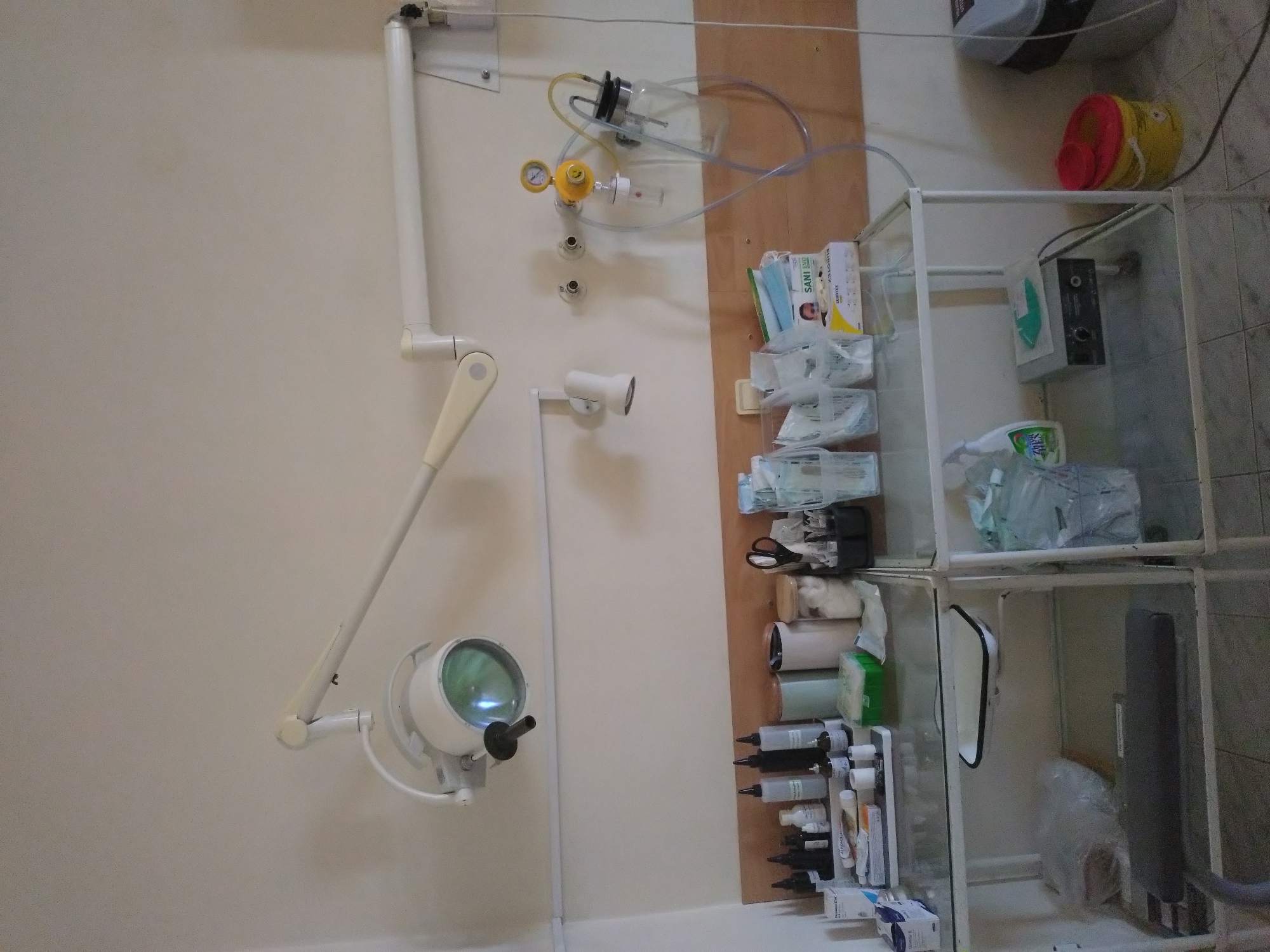 The image shows a medical examination room. On the left is an examination light attached to the wall. There's a gas delivery system with a pressure gauge and oxygen source above a small spotlight. To the right, a tall, white metal shelving unit displays medical supplies such as masks, syringes, and disinfectants. In the background, the floor is tiled. The location is likely a medical facility in Stara Zagora, Bulgaria.
