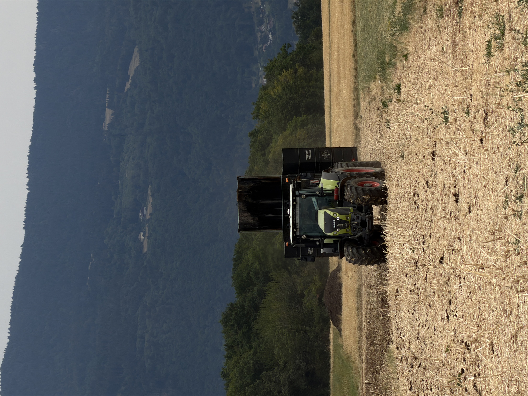 A tractor with a trailer is operating in a field near Reignier-Ésery, France. The tractor is green and black and appears to be hauling material in the black trailer. The field is dry and stubbly, suggesting it may have recently been harvested. In the background, there are trees and a large hill covered in dark green trees. The sky is overcast. The scene appears to be a rural agricultural setting.