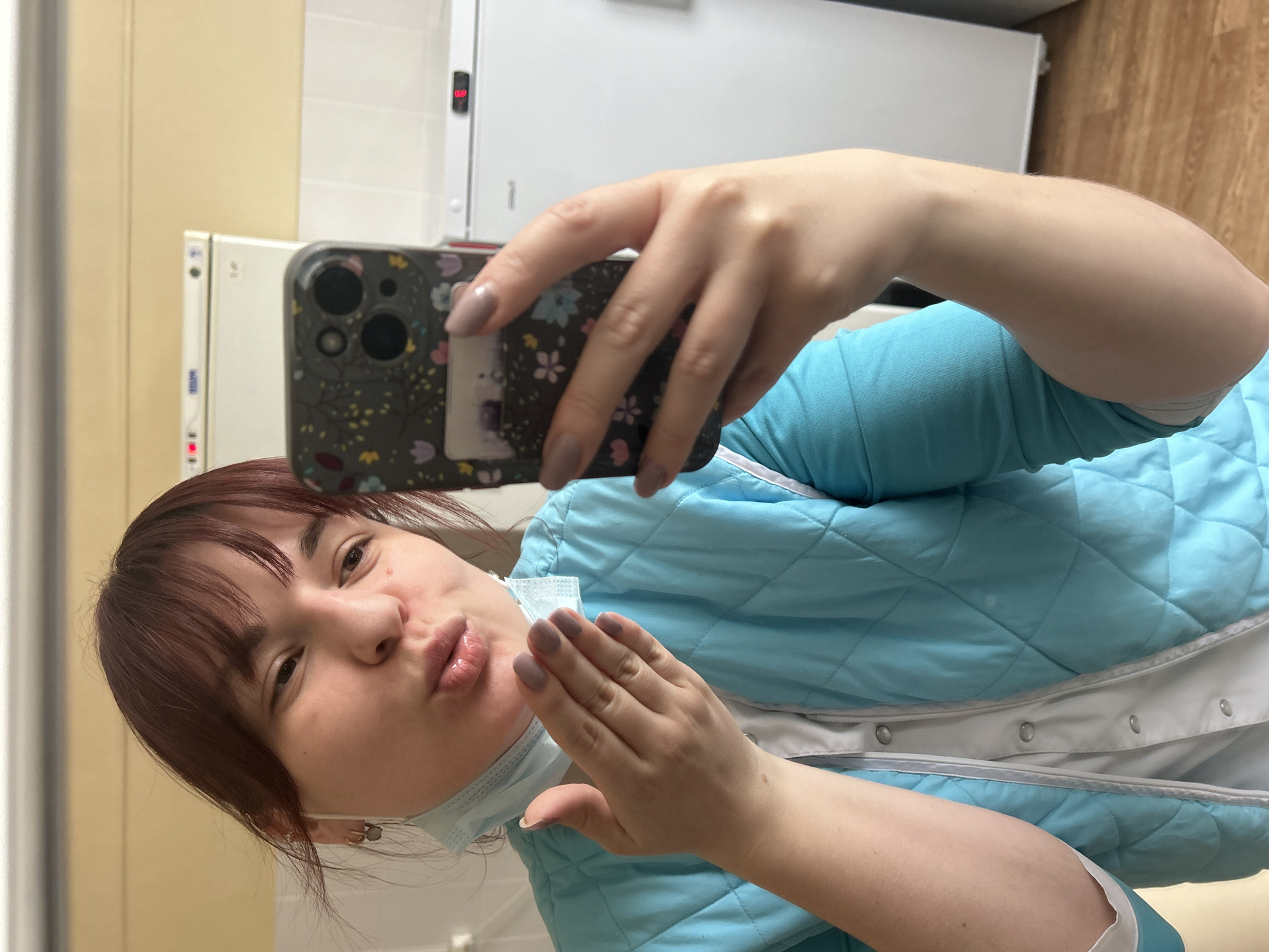 A selfie, taken in what appears to be a mirror, features a woman with reddish-brown hair and bangs. She is captured making a playful "kissing" or "pouty" face directly at the camera. Her right hand holds a smartphone, identifiable by its camera cluster and a floral-patterned case, while her left hand is raised, seemingly in a gesture of blowing a kiss, with fingernails painted a muted grey-purple.
She is dressed in a light blue quilted jacket or vest worn over a white garment, possibly a uniform. A light blue face mask is pulled down, resting around her chin.
The background consists of plain walls, with a light beige section on the left and a white section on the right, typical of an indoor setting. Two large, white industrial-style appliances, likely refrigerators or freezers, are prominent behind her. One features a small red digital display showing numbers such as "1.9" or "1.0", and another display further right shows "8.9" or "8.0", suggesting temperature readouts. A wooden floor is visible in the upper right corner of the frame. The uniform, mask, and industrial equipment strongly suggest a professional or institutional environment, such as a clinic, laboratory, or a facility in Фоки, Russia, where temperature control is essential. No discernible text is clearly visible on any objects, other than the unreadable numbers on the appliance displays. The image conveys a moment of lightheartedness in a functional setting.
