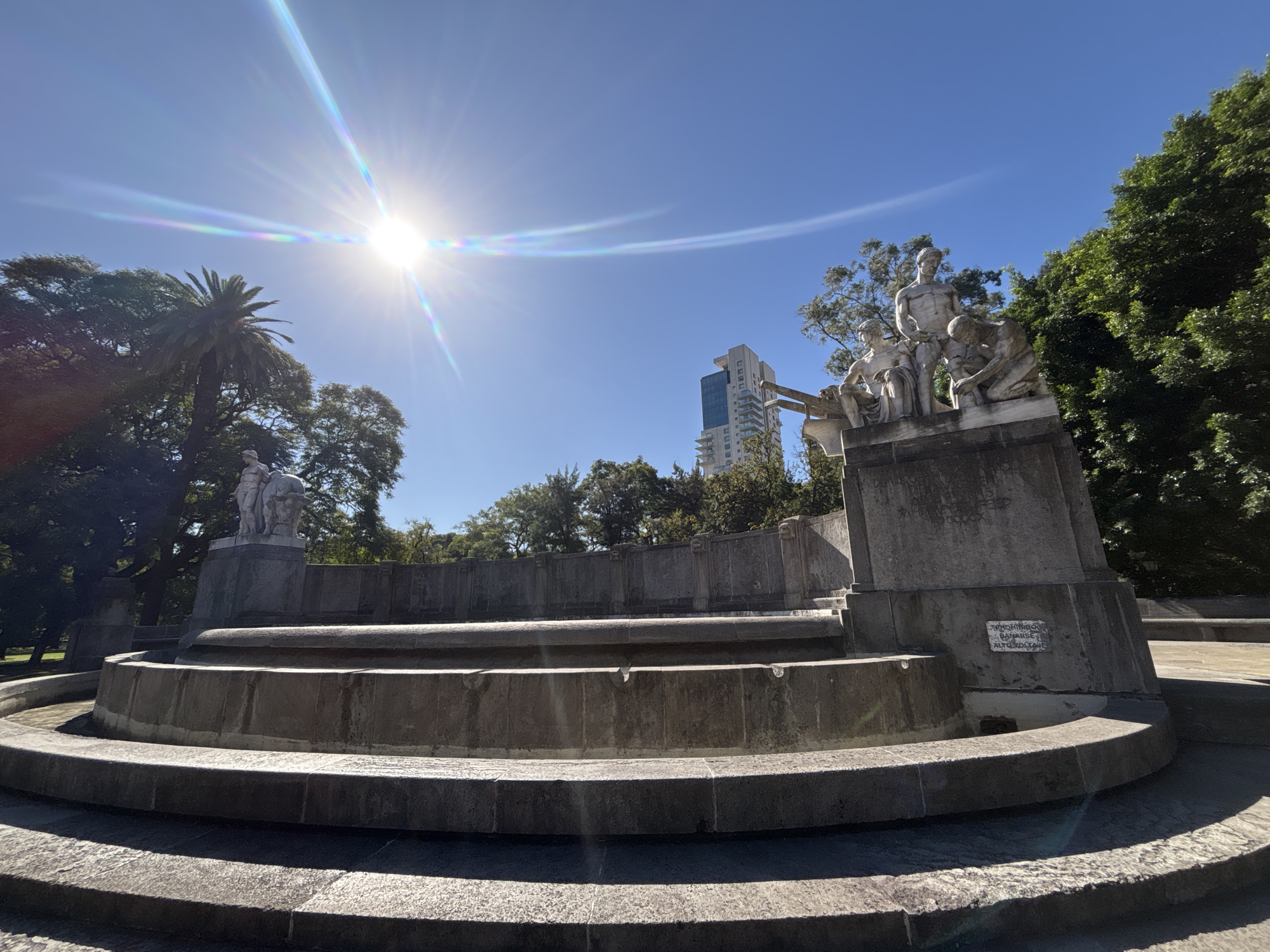 This wide-angle photograph captures a grand, weathered stone monument and multi-tiered fountain under a bright, clear blue sky. The camera's low angle looks up at the monument, centered around a series of circular or semicircular stone steps and basin structures that form the fountain.
On the elevated platforms of the monument, several classical-style white stone statues are visible. On the right, a group of three male figures is depicted: one stands prominently, another is seated, and a third kneels. A less distinct, solitary white statue is also visible on a platform to the far left.
Lush green foliage, including a tall palm tree on the left, surrounds the monument, suggesting a park or public square setting. In the background, a modern high-rise building with glass panels contrasts with the historical monument.
The sun is intensely bright in the upper left, creating a prominent lens flare that streaks across the sky, indicating a sunny, clear day, likely around midday.
On a white plaque affixed to the right side of the monument's base, the text "P. ROCHE BLANCHE", "H. MENARDET", and "AUTORES" is visible, identifying the creators. Carved into the stone above the plaque, partial inscriptions include "PROVINCIA DE", "EL PUEBLO ARGENTINO A", and "MCMX" (1910), suggesting a dedication from the Argentine people or a province, and a creation date.
The monument shows signs of age and weathering, with visible textures and some wear on the stone surfaces. The setting is an urban park in Buenos Aires, Argentina.
