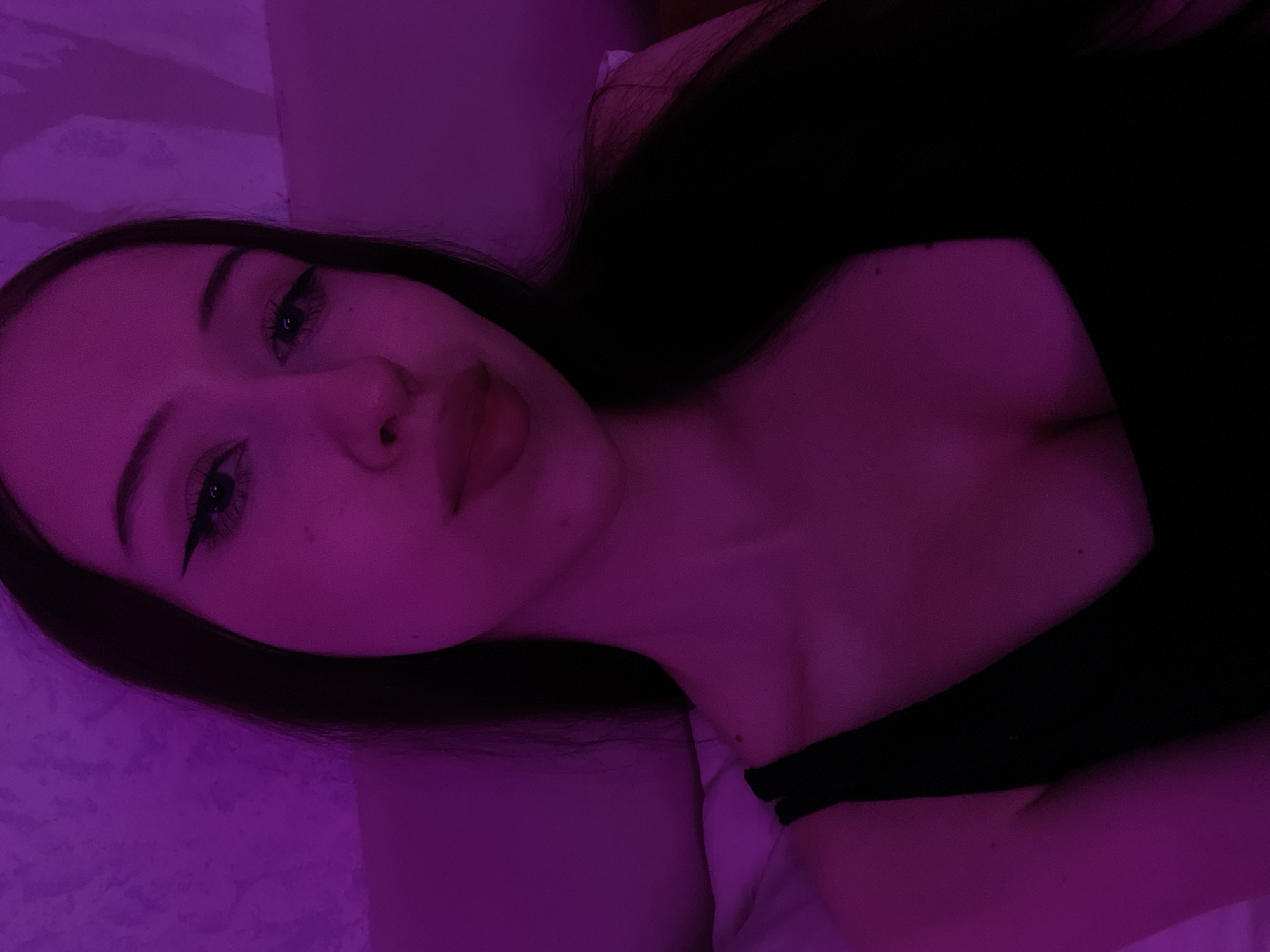 The image is a close-up, selfie-style photograph of a young woman with long, dark hair and fair skin. She is illuminated by a purple light, which casts a strong hue over the scene. Her makeup includes winged eyeliner and lip gloss. She is wearing a black, low-cut top. The background appears to be a textured wall with a subtle pattern. There are no discernable landmarks or elements that would place this image specifically in Votkinsk, Russia.