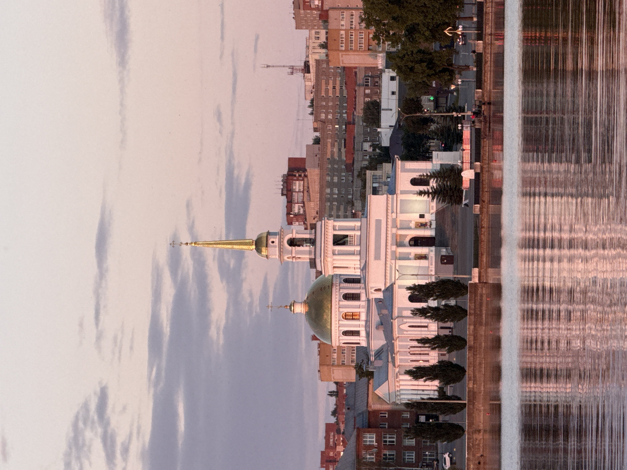The image depicts the city of Votkinsk, Russia, at either sunrise or sunset, centered on the Transfiguration Cathedral (Spaso-Preobrazhensky Cathedral) situated on the bank of the Votkinsk Pond. The sky displays hues of soft pink, orange, and light blue, with scattered light grey clouds.
The Transfiguration Cathedral, a prominent white neoclassical structure, occupies the midground. It features two golden domes: a larger, central onion-shaped dome topped with an Orthodox cross, and a taller, slender golden spire on a bell tower to its right, also surmounted by a cross. The cathedral's facade includes multiple arched windows and entrances, some of which show an internal amber glow, indicating illumination. Columns are visible on the bell tower and surrounding the main dome's base.
Behind the cathedral, a dense urban landscape consists of multi-story buildings, varying in age and style. Some are older, reddish-brown brick structures, while others are grey or beige block-style residential or administrative buildings. A tall communication antenna is visible on a rooftop in the distant right.
In the foreground, the calm surface of the Votkinsk Pond reflects the sky's colors and the cathedral's illuminated structure, creating elongated, shimmering vertical streaks. A concrete or stone embankment separates the water from the land, along which a row of dark green coniferous trees (likely cypress or spruce) is planted. Beyond the trees, a paved area or street is visible, with faint streetlights and a partially obscured white car on the far left. On the right side of the embankment, faint figures or objects are discernible near the water's edge.