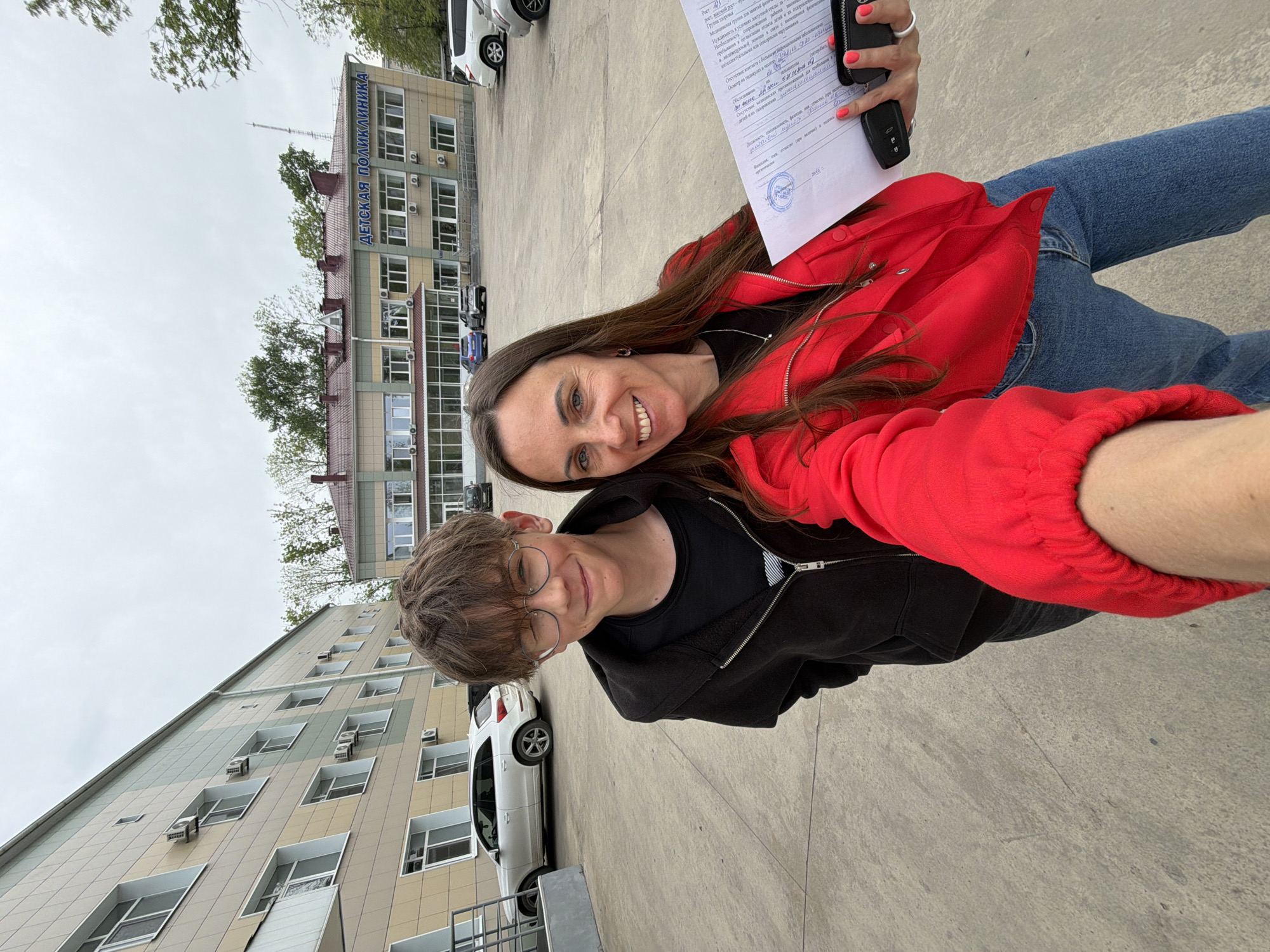 Two individuals, a younger person wearing glasses and a black hoodie, and an older person wearing a red jacket and jeans, are posing for a selfie. They are standing in a paved outdoor area, likely a parking lot or plaza, with buildings visible in the background. The building on the left is a multi-story structure with light-colored tiles and numerous windows, many of which have air conditioning units installed. The building behind them reads "ДЕТСКАЯ ПОЛИКЛИНИКА" (Detskaya Poliklinika), which translates to "Children's Polyclinic." The OCR text in the image suggests the location is Birobidzhan, Russia. The older person is holding a piece of paper and a car key in their hand. Several cars are parked in the vicinity. The sky is overcast, indicating a cloudy day.