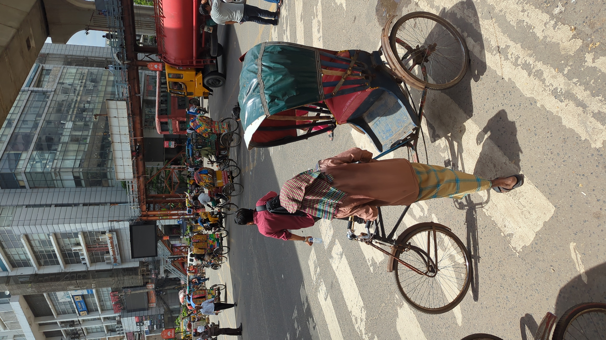 The image depicts a street scene in Dhaka, Bangladesh, likely in a commercial or high-traffic area. In the foreground, a man wearing a brown shirt and a yellow checkered lungi is pushing a rickshaw. He has a checkered scarf draped over his head and shoulders. The rickshaw has a blue and red carriage with a green canopy. A man in a maroon shirt walks away from the viewer, holding a bottle of water, and a black backpack is slung over his shoulder. In the mid-ground, several other rickshaws, some laden with goods, are present on the road. Pedestrians are visible, and traffic appears to be present, including a yellow truck with a red tank, and a red bus. Buildings line the background, with modern architecture characterized by glass facades, suggesting a developed urban environment. An elevated road or flyover is also visible in the background. The scene is illuminated by sunlight, casting shadows on the road, which is marked with a pedestrian crossing.