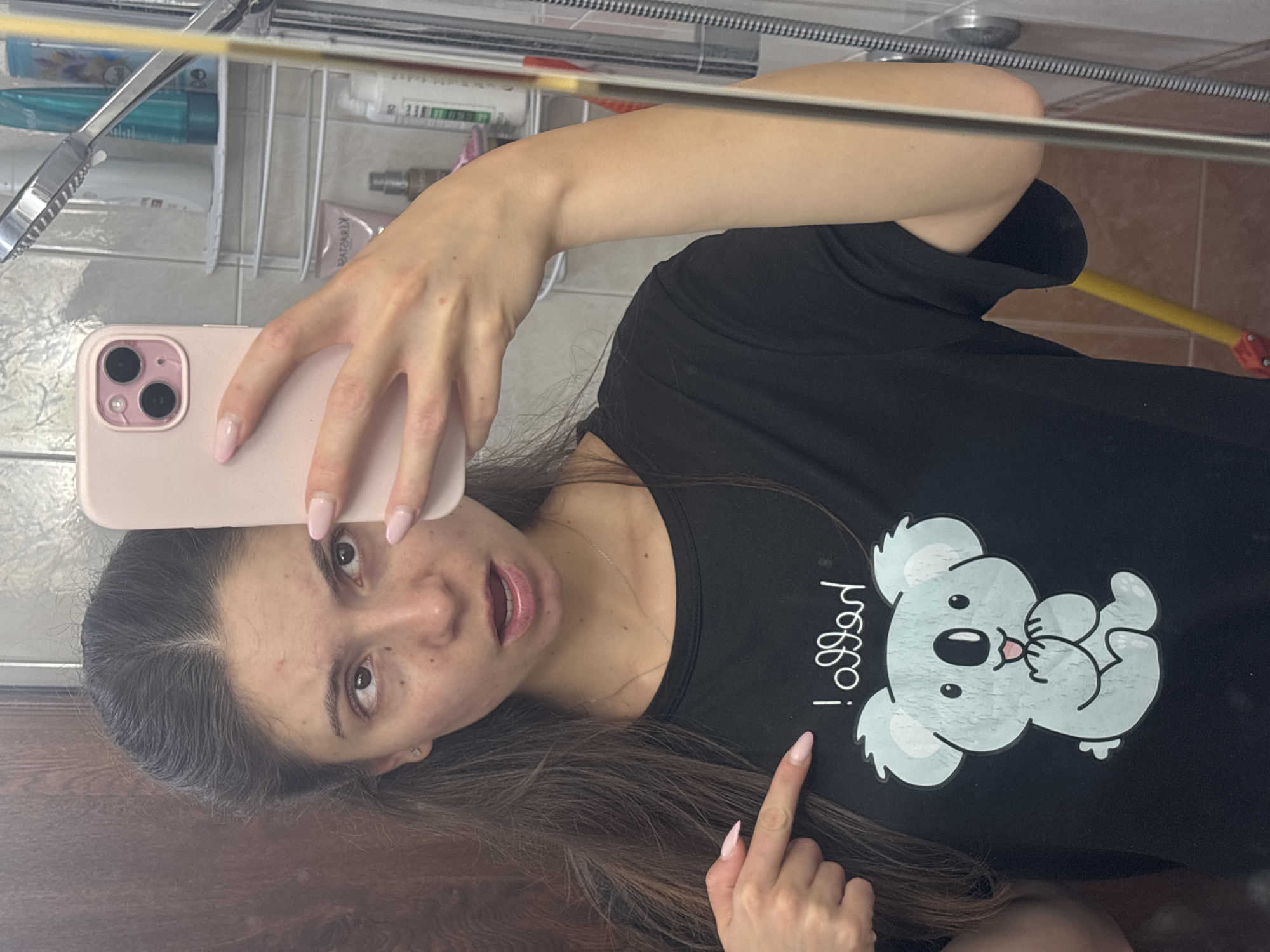 A young woman is taking a mirror selfie in a bathroom. She is wearing a black t-shirt with a cartoon koala and the word "hello" printed on it. She is pointing at the t-shirt with her left hand. She is holding a pink smartphone in her right hand to take the picture. The bathroom appears to be tiled and has a shower visible in the background. The woman has long, dark hair and is looking at the camera with her mouth slightly open. She has light skin and appears to be in her late teens or early twenties. The scene is domestic and private, with no indication of the specific location within Varna, Bulgaria.