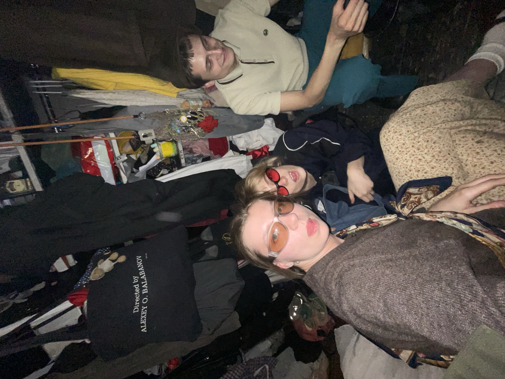 A high-angle, low-light photograph shows three individuals amidst a cluttered indoor space, likely a living area, with assorted objects scattered around.
A young man with short brown hair, wearing a light-colored collared shirt and blue pants, lies on his back and is smiling. Another individual with fair hair and red sunglasses lies closer to the foreground, nestled among various items of clothing and other objects. The third person, sporting orange-tinted glasses, is partially visible and lying nearby in a brown sweater.
The surrounding area contains clothing items in black, white, and yellow, as well as a shopping bag. A black pillow or cushion with the text "Directed by ALEXEY O. BALABANOV" is visible among the clothing. The scene is disorganized, with the environment containing a mix of personal belongings.
The photo's low lighting makes it difficult to discern precise details, but there's no information to confirm its location as Moscow.