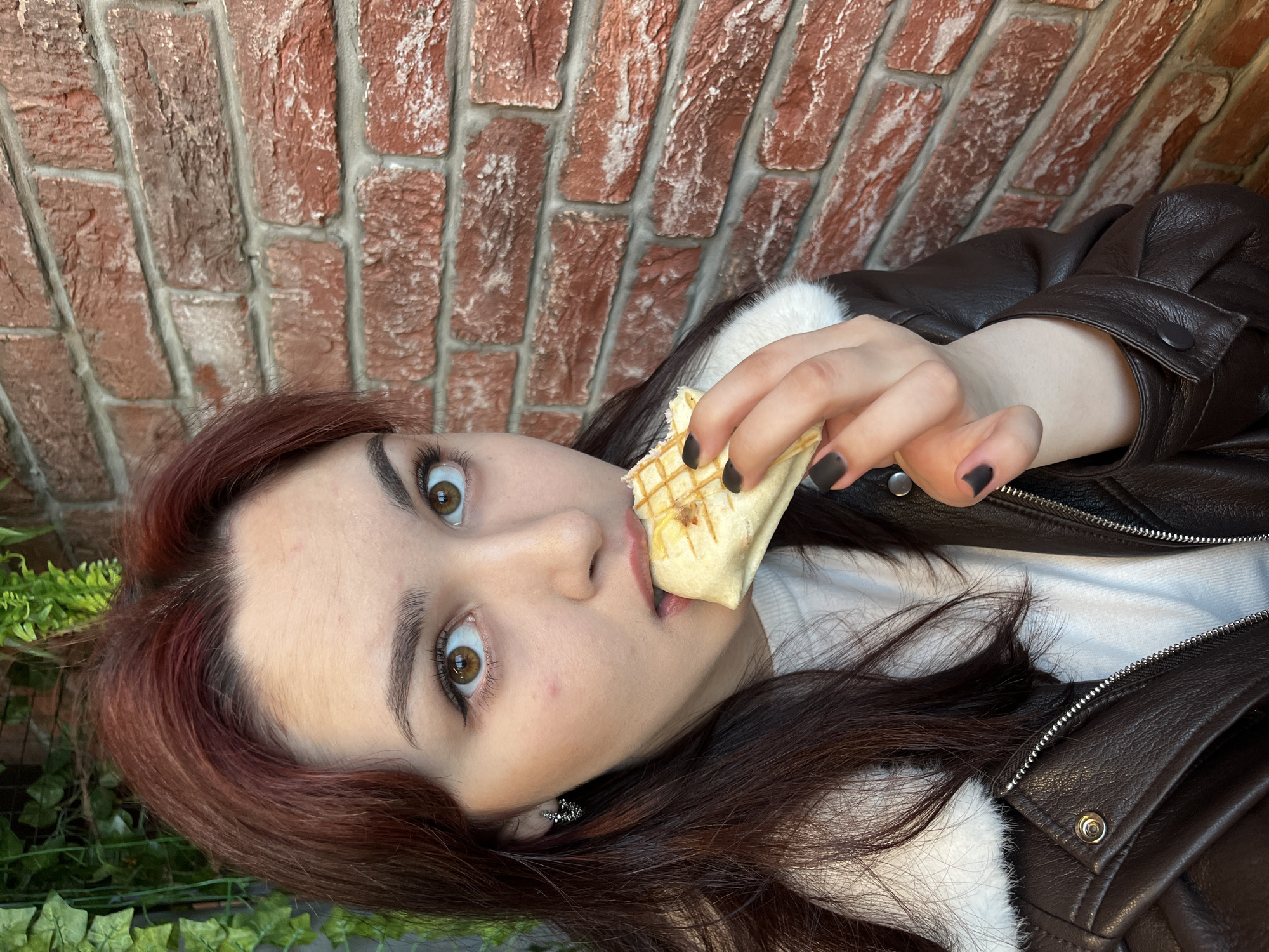 A young woman with auburn hair and brown eyes is shown eating a sandwich. She is wearing a white shirt and a brown leather jacket with a white furry collar. Her nails are painted black. She is positioned in front of a brick wall, and some green foliage is visible at the bottom left of the frame. The location, Neftekamsk, Russia, is based on prior context/metadata, but is not inherently visible in the provided image.