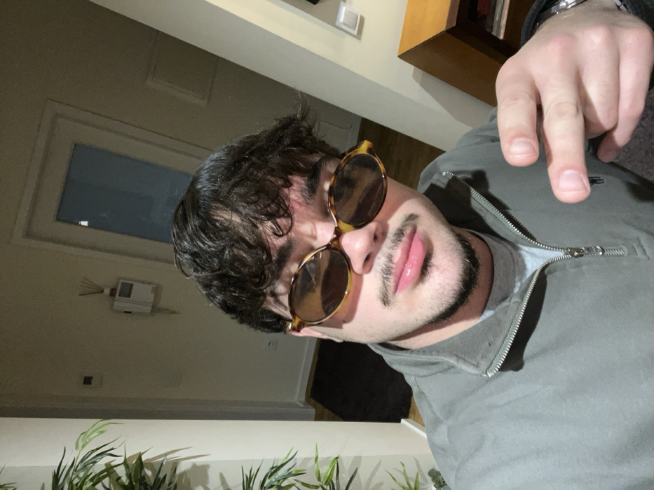 A man with dark, curly hair and a goatee wears round, tortoiseshell sunglasses. He's wearing a gray zip-up jacket and a watch. His hand is raised, forming a gesture. A door and a wall-mounted device are visible in the blurred background, along with a bookcase. The lighting is somewhat dim, creating shadows on the man's face and jacket. A plant is visible in the lower portion of the frame. The image is rotated.
