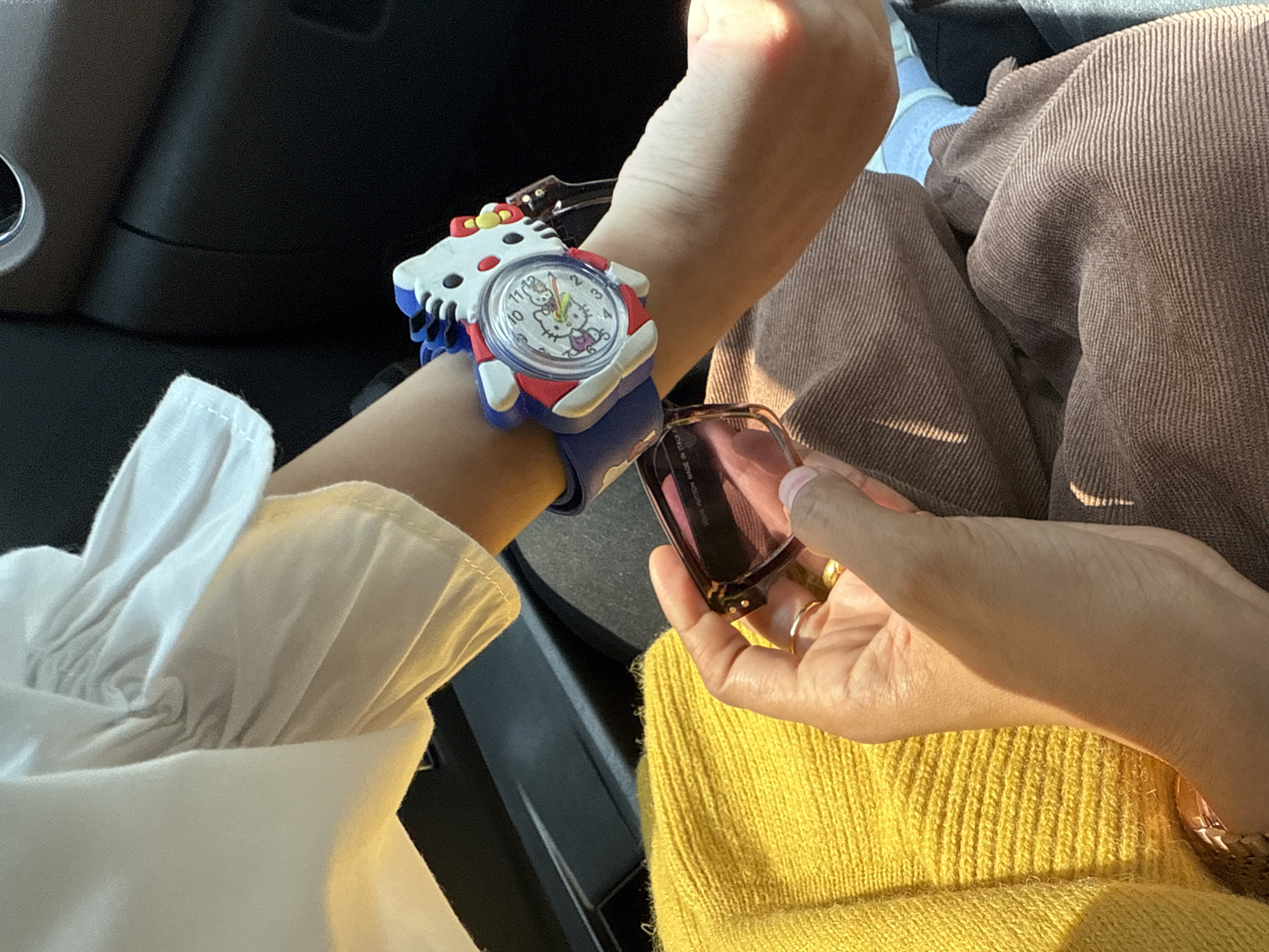 The image shows a close-up of a person's arm and hand, with a Hello Kitty watch on the wrist. The person is wearing a white, ruffled shirt and a yellow sweater. In their hand, they are holding a pair of sunglasses with a tortoiseshell frame. The scene appears to be inside a vehicle, as indicated by the dark interior and a vent visible in the background. The lighting suggests it is daytime. There is no specific information or visual element within the image to suggest a location in Tiga Raja, Indonesia.