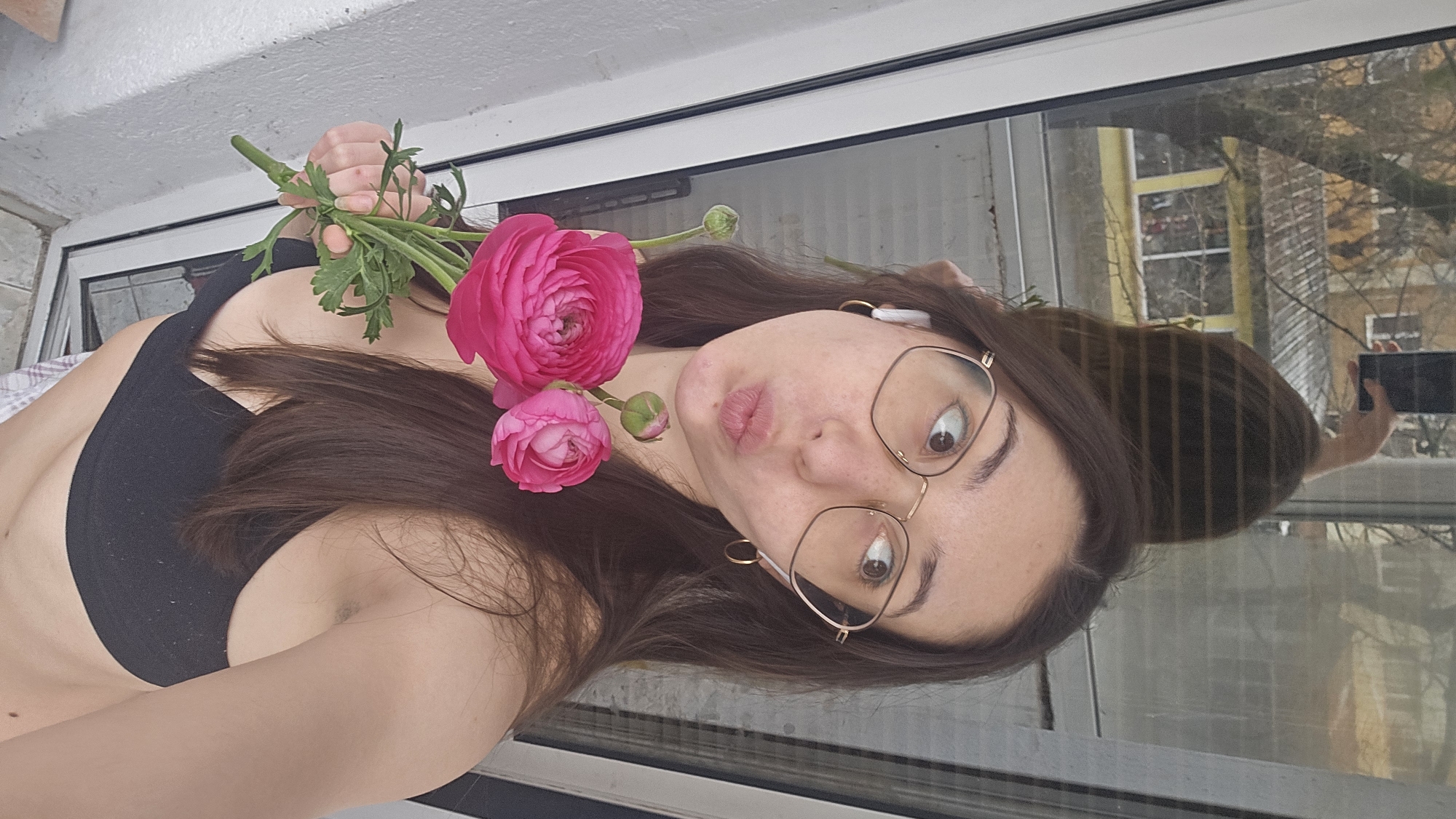 A woman with brown hair and gold-rimmed glasses is holding pink flowers in front of her face, posing for a selfie in front of a window. She is wearing a black sports bra and wireless earbuds. The window reflects a building and trees, suggesting an outdoor view. The specific location within Stara Zagora, Bulgaria, cannot be determined from the given image.