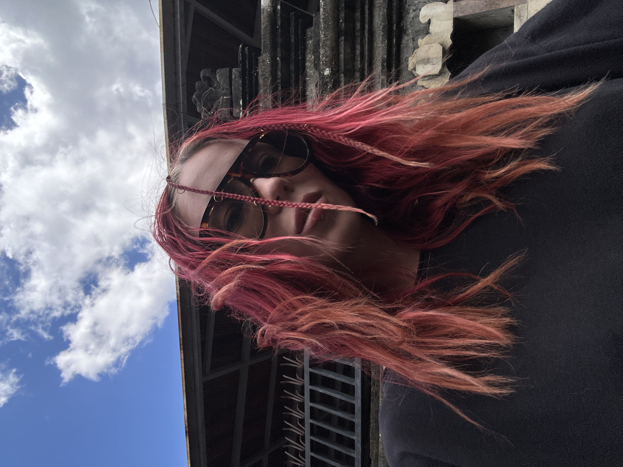 A young woman with brightly dyed pink and red hair, styled in loose waves and two braids adorned with small metal rings, poses for a selfie. She is wearing large, dark-rimmed sunglasses and a black t-shirt. The background features a traditional Balinese-style building with a dark-tiled roof and ornate stone carvings, possibly a temple or an ancient structure. The sky above is a vibrant blue with fluffy white clouds. The setting appears to be outdoors, with some vegetation and architectural elements visible. The overall atmosphere is casual and outdoor.