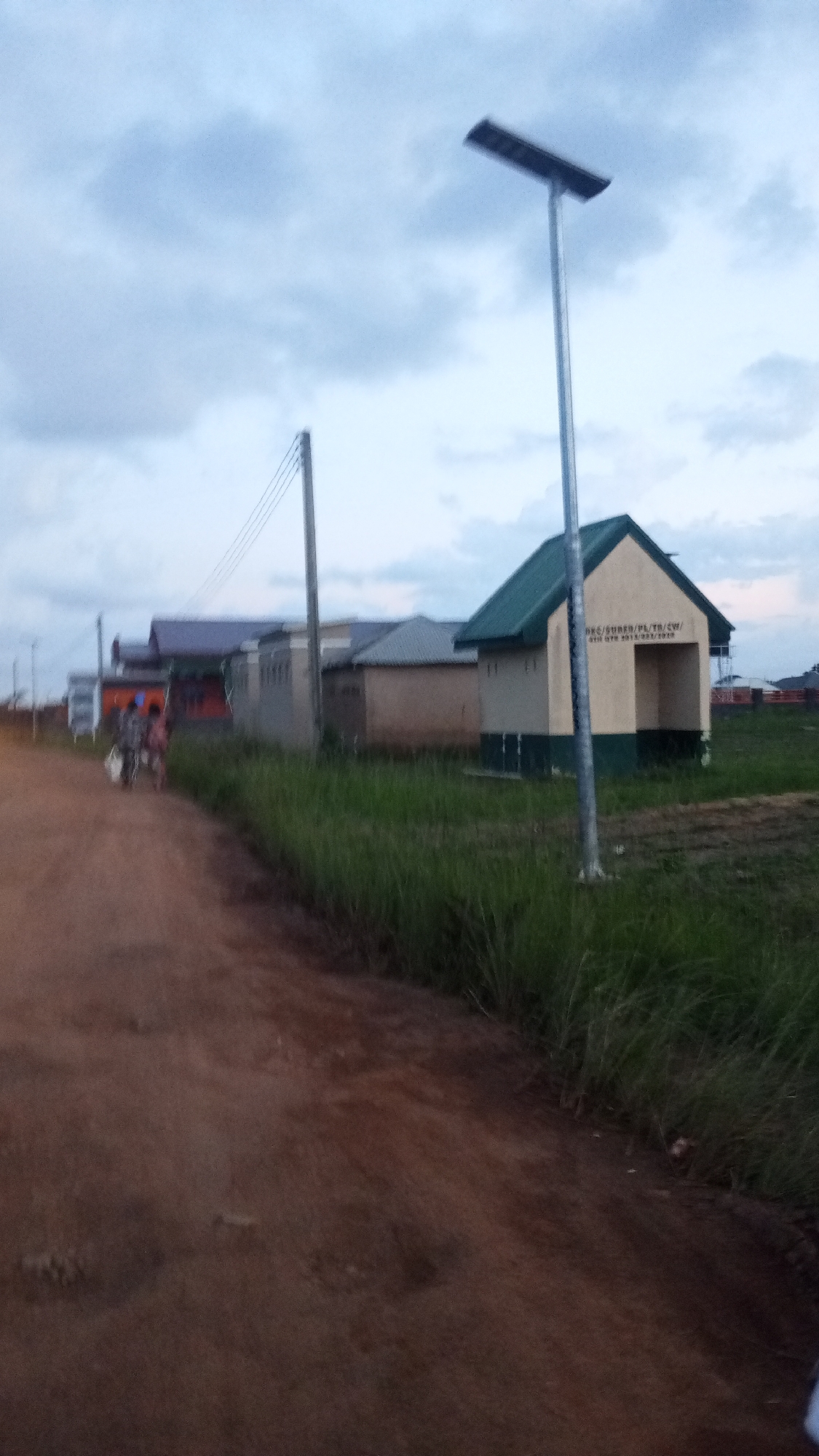 The image depicts a street scene in Kwang, Nigeria, under an overcast sky. A dirt road dominates the foreground, curving to the left. On the right side of the road, tall, green grass grows. In the background, a building with a green roof and beige walls stands out, featuring an inscription that appears to read "DEC/SUBEB/PL/TB/CW/ 6TH OTR 2013/2017/2018". A solar-powered street lamp is visible behind this building, its panel angled towards the sky. To the left of this building are other structures with varying roof colors and designs. A utility pole with electrical wires is also present. In the middle ground, on the dirt road, two individuals are walking away from the camera, carrying bags. The overall atmosphere is subdued, possibly due to the cloudy weather and the late hour.