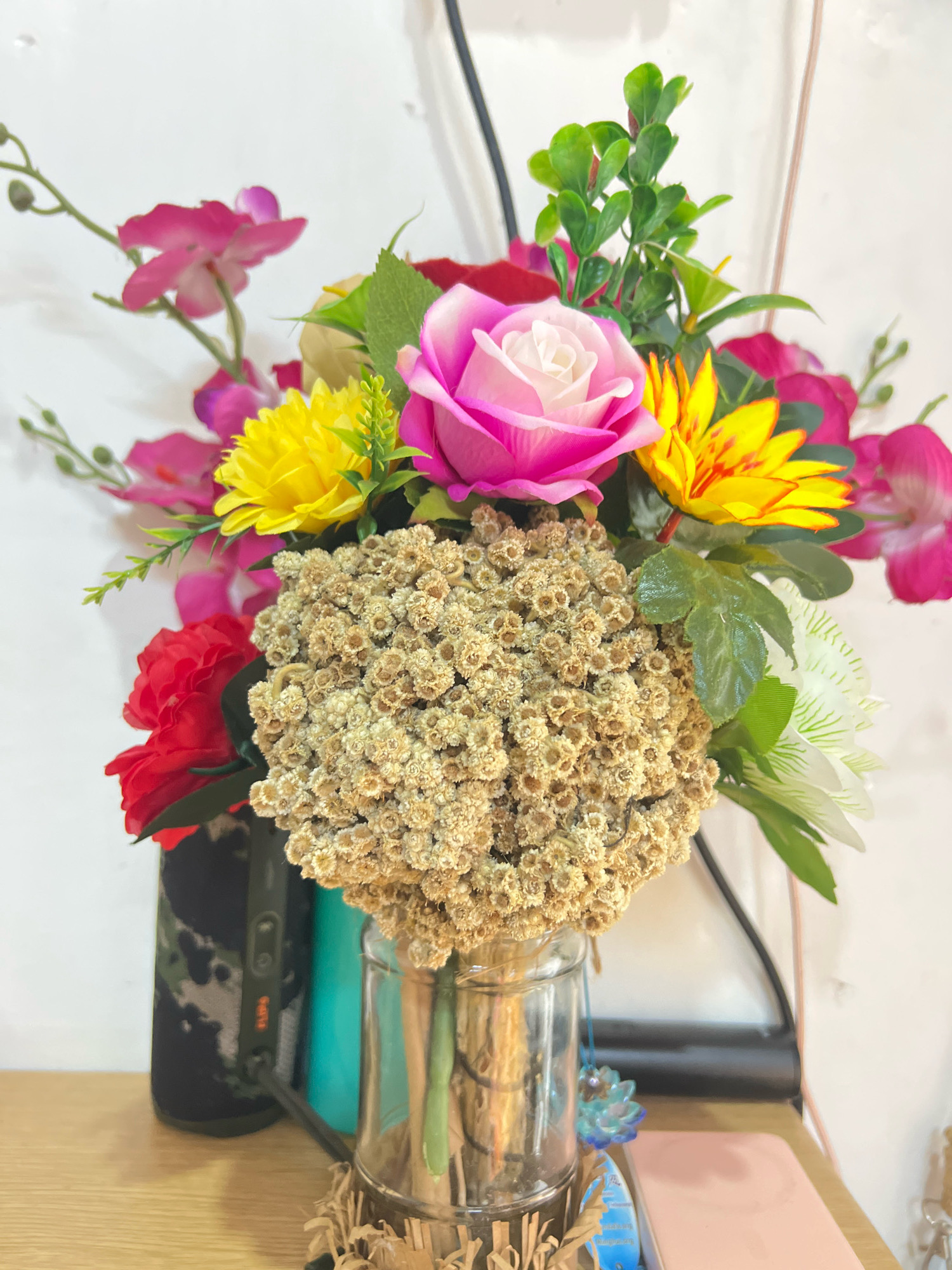 A close-up, indoor, vertical shot showcases a vibrant arrangement of artificial flowers in a glass vase on a wooden surface. The bouquet is a medley of colors and textures, featuring a large pink and white rose as the focal point, surrounded by bright yellow flowers with reddish centers, delicate pink orchids, and a single red rose. A cluster of small, dried, tan flowers forms a dense sphere in the center of the arrangement. Green foliage is interspersed throughout, adding depth and contrast. The vase, made of clear glass with a metallic rim, is partially filled with water. Behind the flowers, a white wall serves as the backdrop. To the left of the vase, a dark object with a camouflage pattern and a teal cylindrical object are visible. The overall scene suggests a decorative display in a home or office. There are no people or animals present, and no actions or interactions are depicted. The image does not provide any specific information about its location in Denpasar, Indonesia.