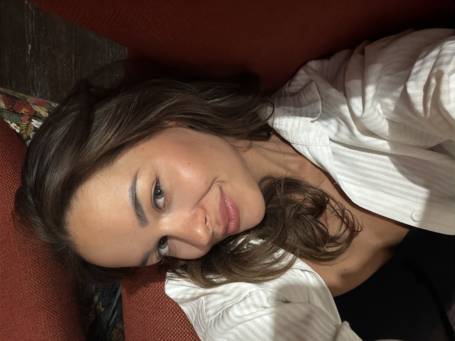 The image displays a close-up portrait of an adult female, captured from a slightly high angle, consistent with a selfie. She has brown, wavy hair that falls around her shoulders and light-colored eyes. Her skin exhibits a smooth texture, and her lips are tinted with a light pink hue, curving into a subtle smile as she looks directly at the camera. The subject is dressed in a white, collared shirt featuring faint vertical stripes, worn unbuttoned over a dark, solid-colored inner garment. A delicate, silver-toned chain necklace is visible around her neck. She is leaning against a reddish-orange upholstered surface, indicative of a sofa or armchair. To the upper left, a small portion of a multi-colored fabric with a floral pattern is visible, suggesting an adjacent cushion or upholstery element. A dark, textured surface, possibly wood, forms the background behind her. No visual information within the image allows for the identification of a specific location within the city of Krasnoyarsk, Russia.