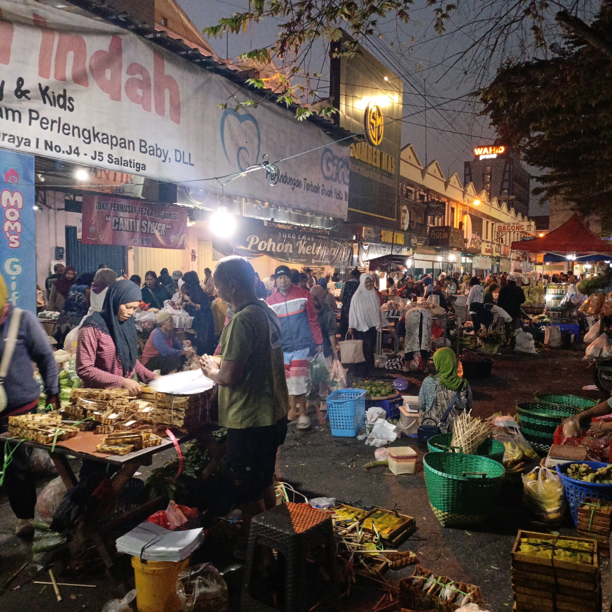 This is a medium-shot, eye-level photograph taken at dusk or early evening, depicting a bustling outdoor market scene in Salatiga, Indonesia. The image is focused on a vendor stall where a woman wearing a black hijab and a striped pink and black long-sleeved shirt is arranging goods on a table. In front of her, several stacks of small, intricately woven wooden boxes are laid out, some tied with green string. To her right, a person in a green shirt is also engaged with the merchandise.
The background reveals a crowded market atmosphere with numerous people, many wearing hijabs, browsing or working. Various stalls and shops line the street, some with illuminated signs. Notable signs include "SUMBER MAS" with a golden circular logo, and further down, a red neon sign that reads "WAH'D." The market appears to be set up along a street with overhead wires and branches of trees visible against the twilight sky. The ground is paved and somewhat uneven, with scattered debris and various market items like baskets, bags, and produce.
The overall impression is one of a vibrant, active marketplace characteristic of Indonesian city life, specifically located in Salatiga.