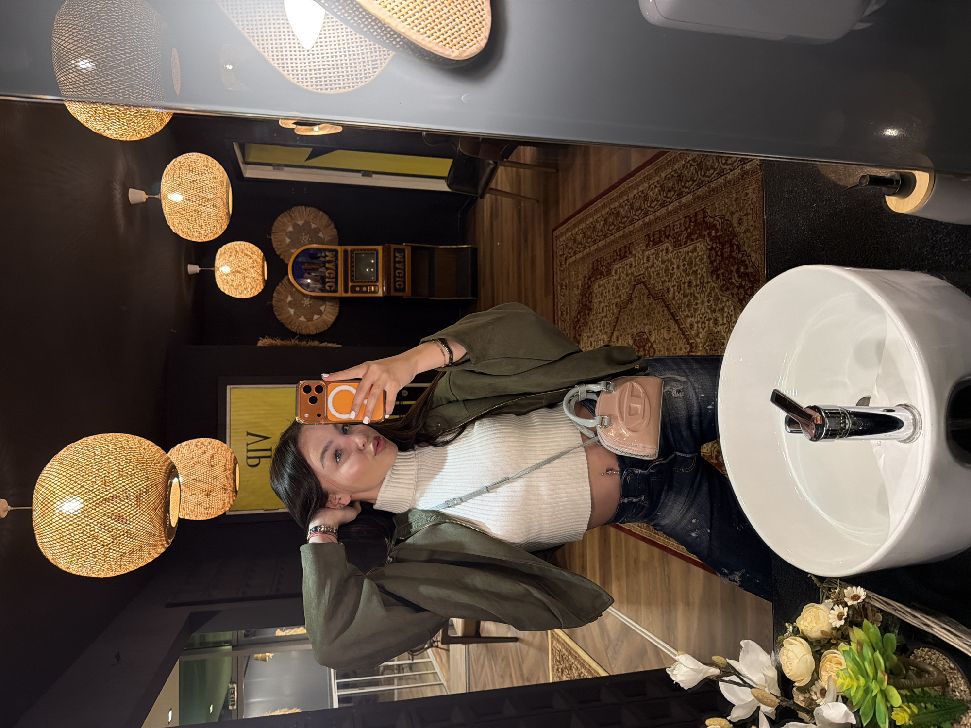 A person is taking a selfie in a bathroom setting, likely in Kulata, Bulgaria, given the prompt. The individual, presumably female, is wearing a white turtleneck sweater, a green jacket, and dark-wash jeans. A small, light pink purse with a strap is slung across their torso. They are holding an orange cell phone in their right hand, taking the photograph in a mirror. The background reveals elements of the bathroom, including a white, circular sink with a chrome faucet and a floral arrangement at its base. The lighting consists of multiple woven, globe-shaped pendant lamps. A vintage-style machine with buttons and a screen is visible, and a patterned rug is on the floor.