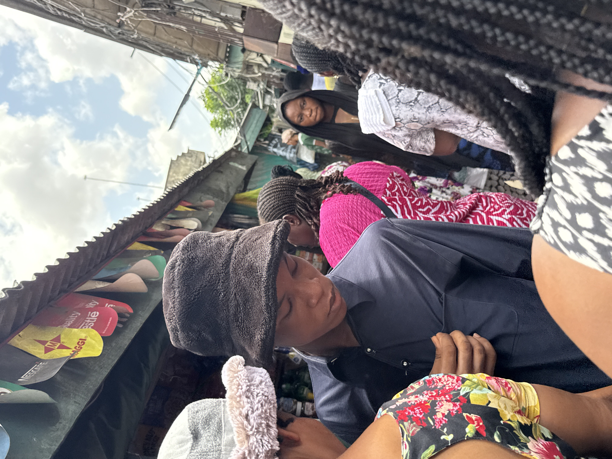 The image depicts a bustling outdoor market scene, characteristic of a vibrant urban environment like Lekki, Nigeria. The perspective is from within a crowd, looking towards market stalls and other people.
In the foreground, the shoulder and braided hair of one individual are visible on the right. To their left, a person wearing a dark blue shirt and a textured, dark grey hooded garment is prominently featured, looking downwards with a focused expression. Their left hand rests near their stomach. Adjacent to this person, part of another individual wearing a black top with a colorful floral pattern is visible.
Further into the midground, several other people are present. A woman wearing a black head covering and a dark top is seen looking directly towards the viewer. Behind the foreground figures, another person in a bright pink and white patterned outfit is visible.
The market stalls lining the scene feature corrugated metal roofs. Underneath these stalls, various products are displayed, including what appear to be shoes or sandals. The scene is illuminated by daylight, with a bright, partly cloudy sky visible above the stalls. Numerous overhead wires and cables crisscross against the sky.
Visible text on product packaging includes prominent brand names like "MAGGI," "Nestle," and "NESCAFE," along with the word "Quality." These indicate common consumer goods often found in such markets. The overall atmosphere suggests a busy, active, and crowded daily market.