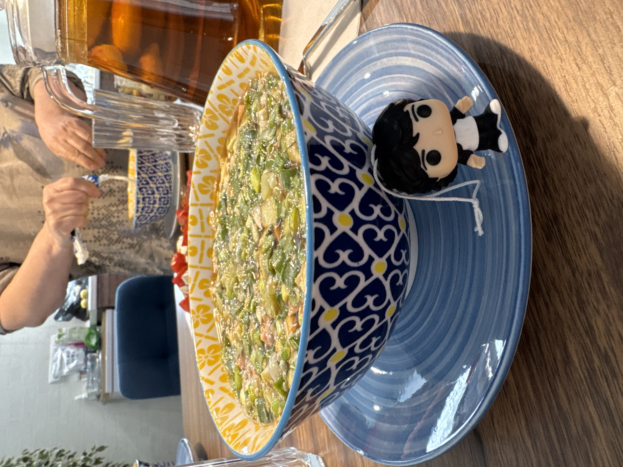 A slightly tilted eye-level shot shows a table setting including a bowl of salad, a plate, a figurine, and a pitcher of tea.
The bowl, filled with a green salad, has a yellow floral pattern on the inside rim and a blue and white heart-shaped design on the outside. It sits on a light blue plate with concentric ridges. A small figurine with black hair and a black and white outfit rests on the plate.
In the background, a large pitcher of what appears to be iced tea is visible. A person's arm is seen reaching for the pitcher with a white object in their other hand. A blue chair is also partially visible in the background.
The table is made of wood and has a warm tone. The lighting suggests the presence of a window or light source to the right of the frame. The environment appears to be a dining area, but specific details about the location in Izhevsk, Russia, cannot be determined from the image.