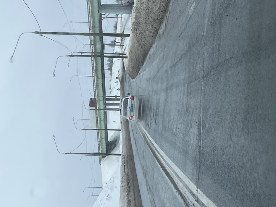 The image depicts a winter or early spring road scene in the city of Ivashevo, Russia, under an overcast sky. A two-lane asphalt road, partially wet with visible puddles and worn double solid white lines, recedes into the distance. A white sedan, identifiable as a police or emergency vehicle by its blue and white checkered roof markings and red light bar, travels away from the viewer in the right lane, leaving a slight water spray from its rear wheels. Its Russian license plate, "P 309 XA 90," is partially visible. Snowbanks, showing signs of melting and dirt, line both sides of the road. Multiple utility poles with streetlights and overhead wires extend along the right side of the road. In the background, an elevated overpass, supported by concrete pillars, crosses above the road. A red semi-truck with a white trailer is observed driving on this overpass. Beyond the overpass, additional snow-covered terrain and sparse, bare trees are visible.