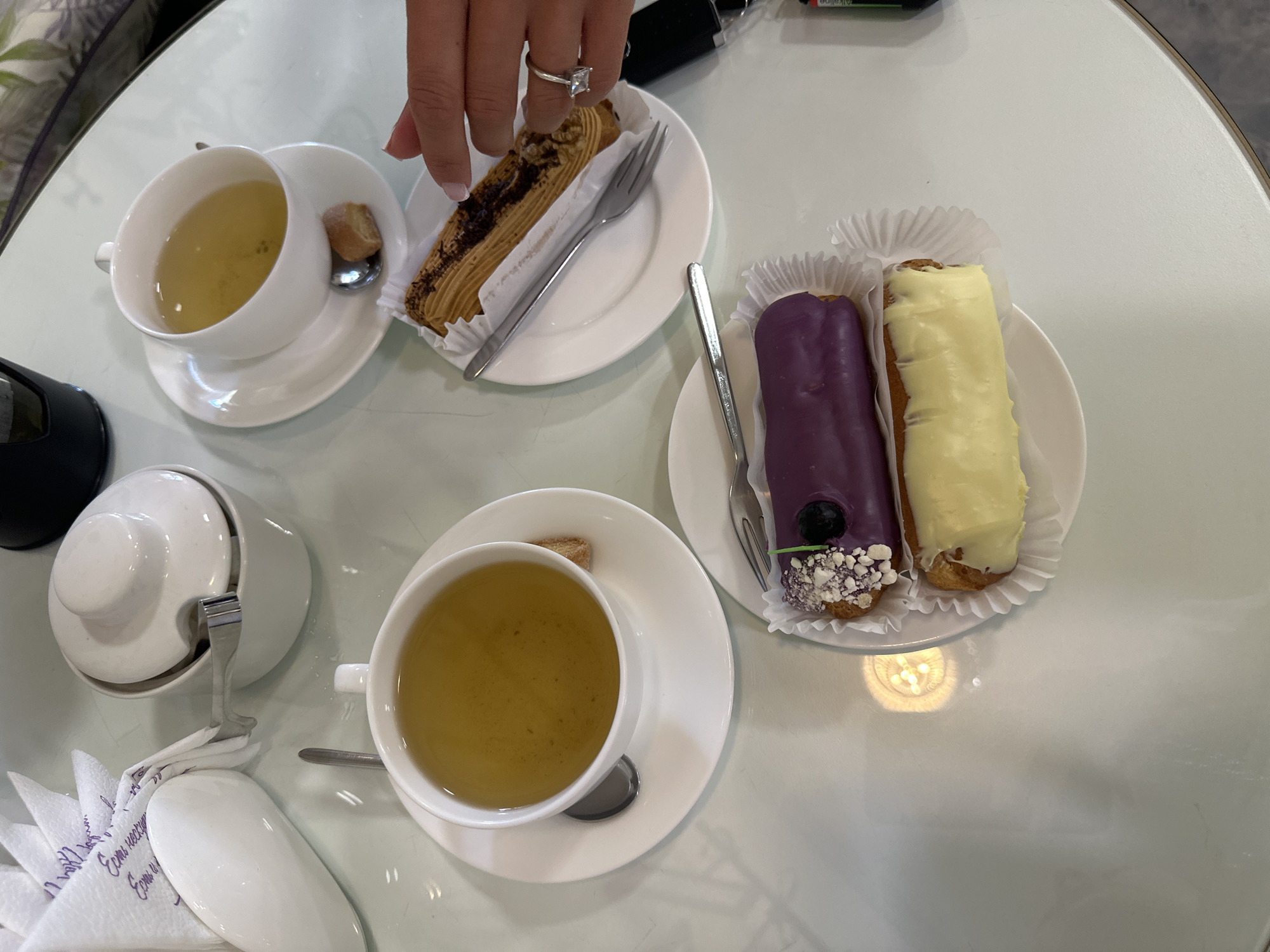 A bird's eye view shows a white, circular table with food and drinks. In the top left corner, a stack of white napkins with purple text is visible. To the right of the napkins is a white sugar bowl with a lid and a silver sugar tong. In the center of the table are two white teacups with saucers, each filled with a light yellow liquid, presumably tea. One teacup has a small cookie on its saucer, and another teacup has a spoon and a small biscuit. In the foreground, two eclairs are presented on a white plate with ruffled paper liners. One eclair is topped with purple icing, white sprinkles, and a dark blue blueberry. The other eclair is covered in yellow icing. A fork rests on the plate next to the eclairs. A person's hand with a diamond ring is visible in the upper right corner, reaching towards an eclair. The scene is likely in a cafe in Perm, Russia, given the context.