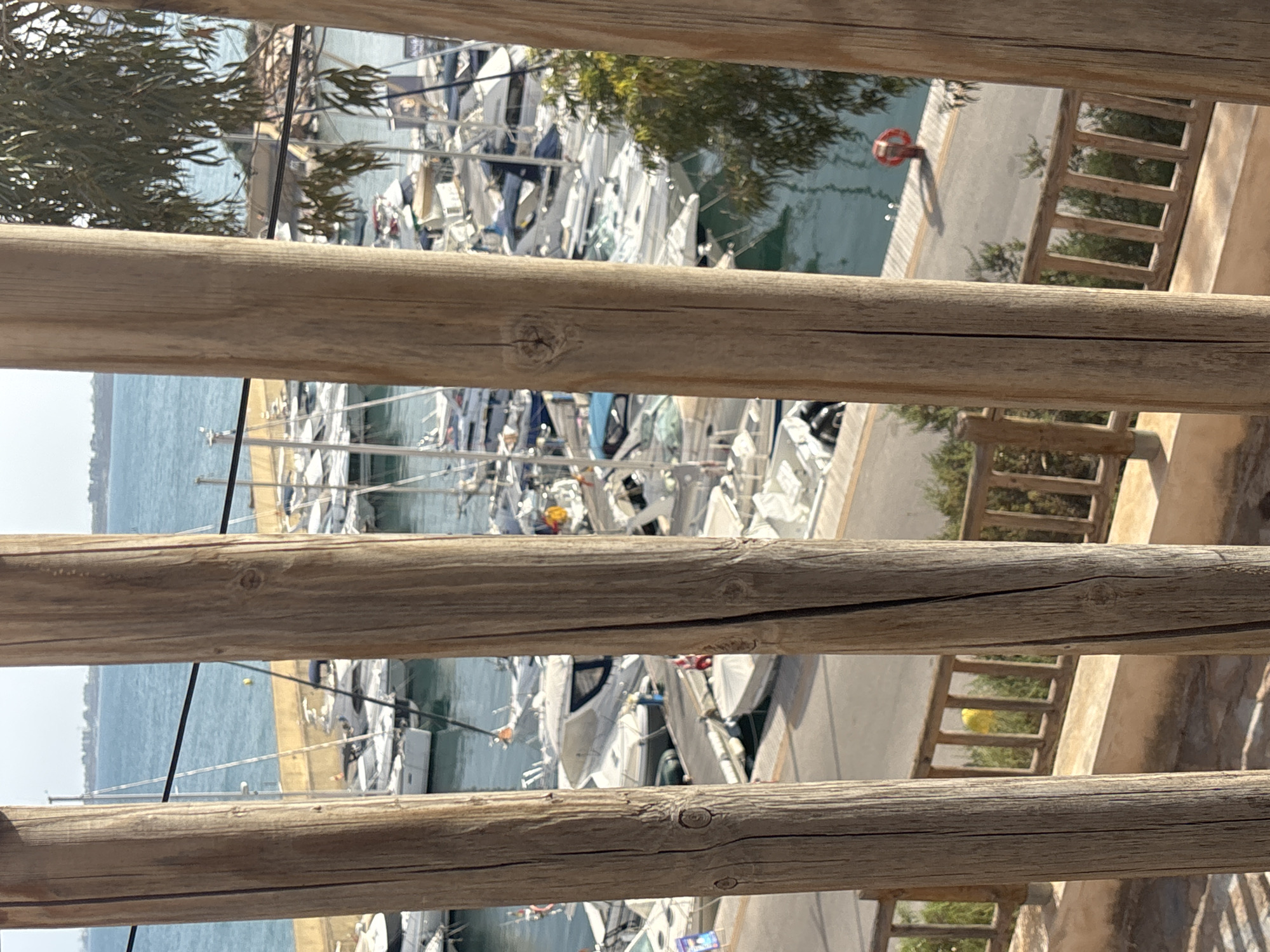 This image depicts a view of a marina filled with numerous boats, primarily sailboats and motor yachts, docked in tranquil blue water. The foreground is dominated by the weathered texture of vertical wooden posts, suggesting a viewpoint from a balcony or deck. Beyond the boats, a paved walkway runs along the waterfront, with a red life preserver visible on the ground. A wooden railing with vertical slats separates the walkway from a slightly elevated area with sparse vegetation and a stone-like surface in the immediate foreground. In the distance, a faint line of buildings and trees hints at the urban landscape of Urbanización Cabo Roig, Spain, under a bright, clear sky.