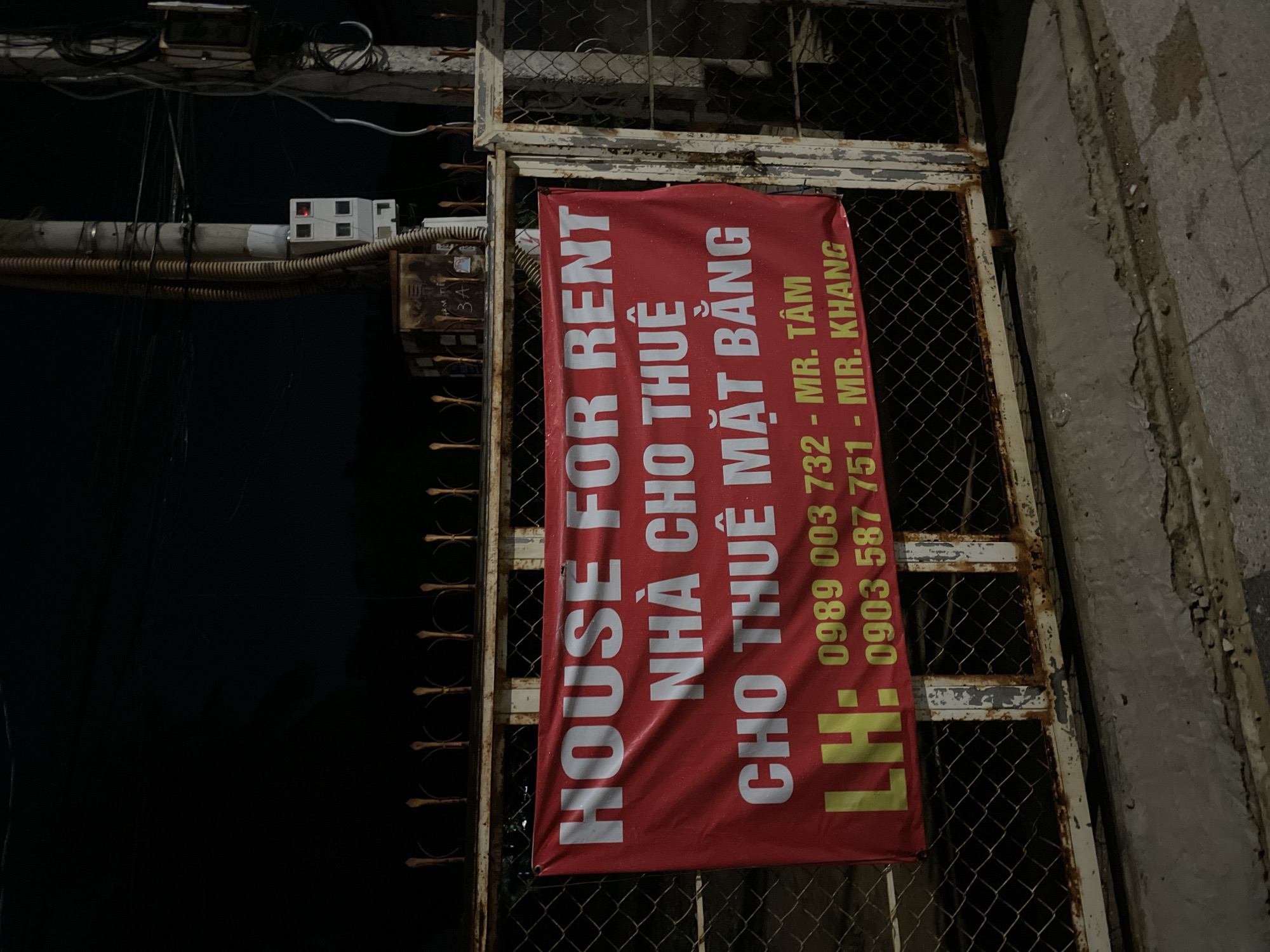 The scene depicts a banner advertising a "HOUSE FOR RENT" and "CHO THUÊ MẶT BẰNG," meaning "SPACE FOR RENT," hanging on a metal gate or fence. The banner is predominantly red with white text in both English and Vietnamese. Contact information, including phone numbers and names (MR. TÂM and MR. KHANG), is provided. The surrounding environment includes electrical wiring and a concrete wall. The location is likely in a Vietnamese neighborhood, given the Vietnamese text and phone numbers. Phu Thuy Ward is a potential location based on the information.