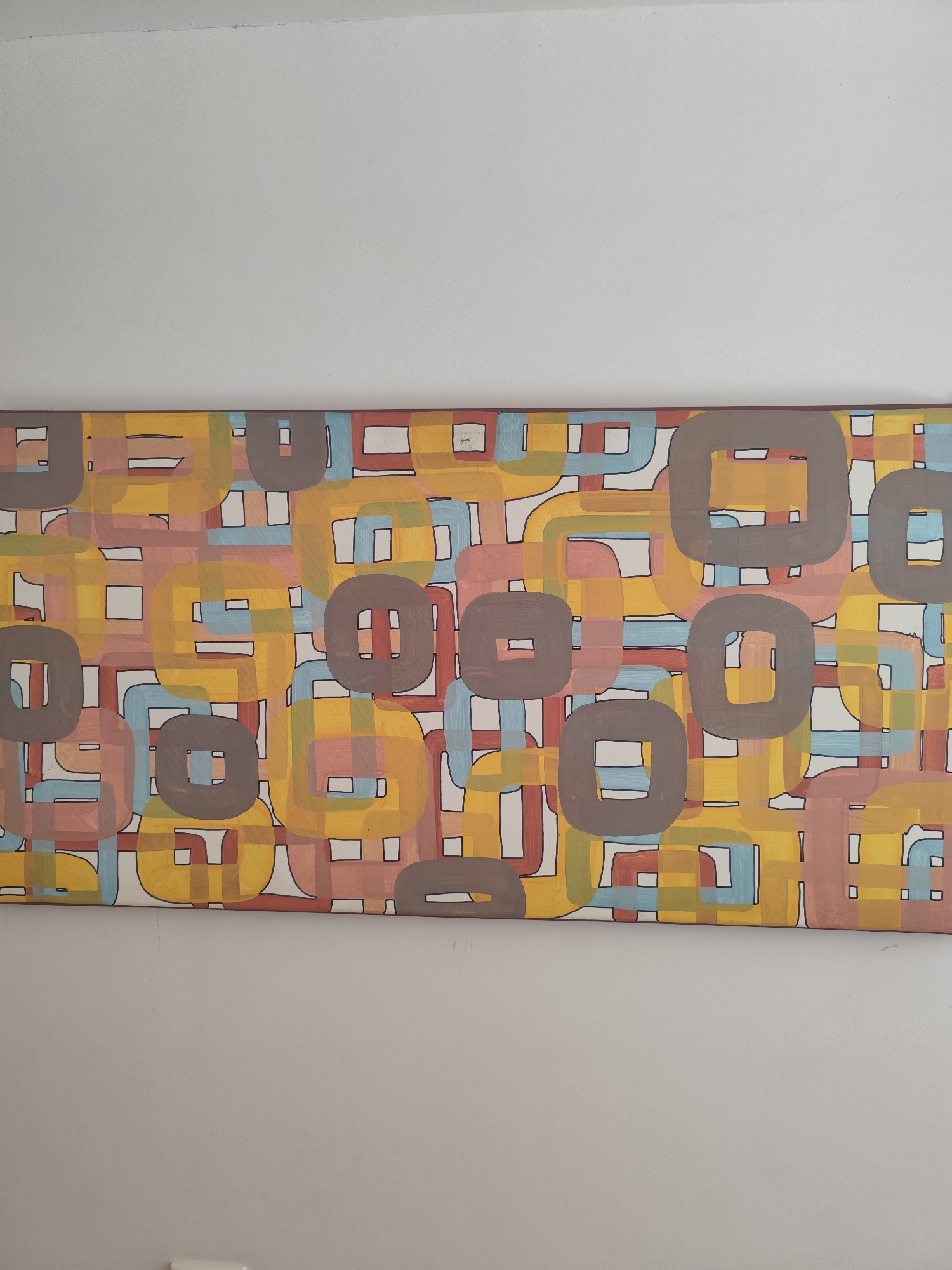 A horizontal, abstract painting hangs on a plain, light-colored wall. The painting features a repeating pattern of rounded squares and rectangular shapes in various colors including yellow, light brown, light blue, and a dusty pink. These shapes are outlined in black and overlap each other, creating a sense of depth and visual complexity. The background of the painting is white. The painting is framed by a thin, dark border. The overall impression is one of a modern, geometric design. The location is unknown.