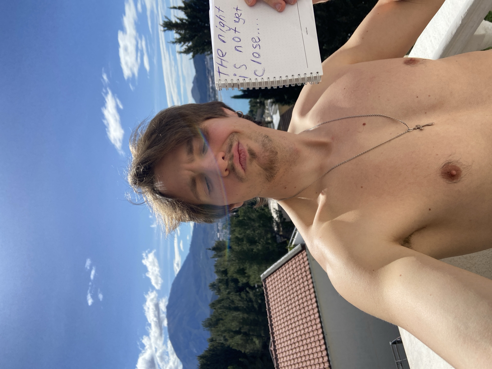 A shirtless man with brown hair, a short beard, and a silver necklace is holding a notepad with the text "The night is not yet close..." written on it. He is outdoors on a balcony or terrace with a red-tiled roof visible below. The background features a mountain range, trees, a body of water, and a bright blue sky with scattered clouds. The scene appears to be in Šušanj, Montenegro, based on the landscape and architectural features.