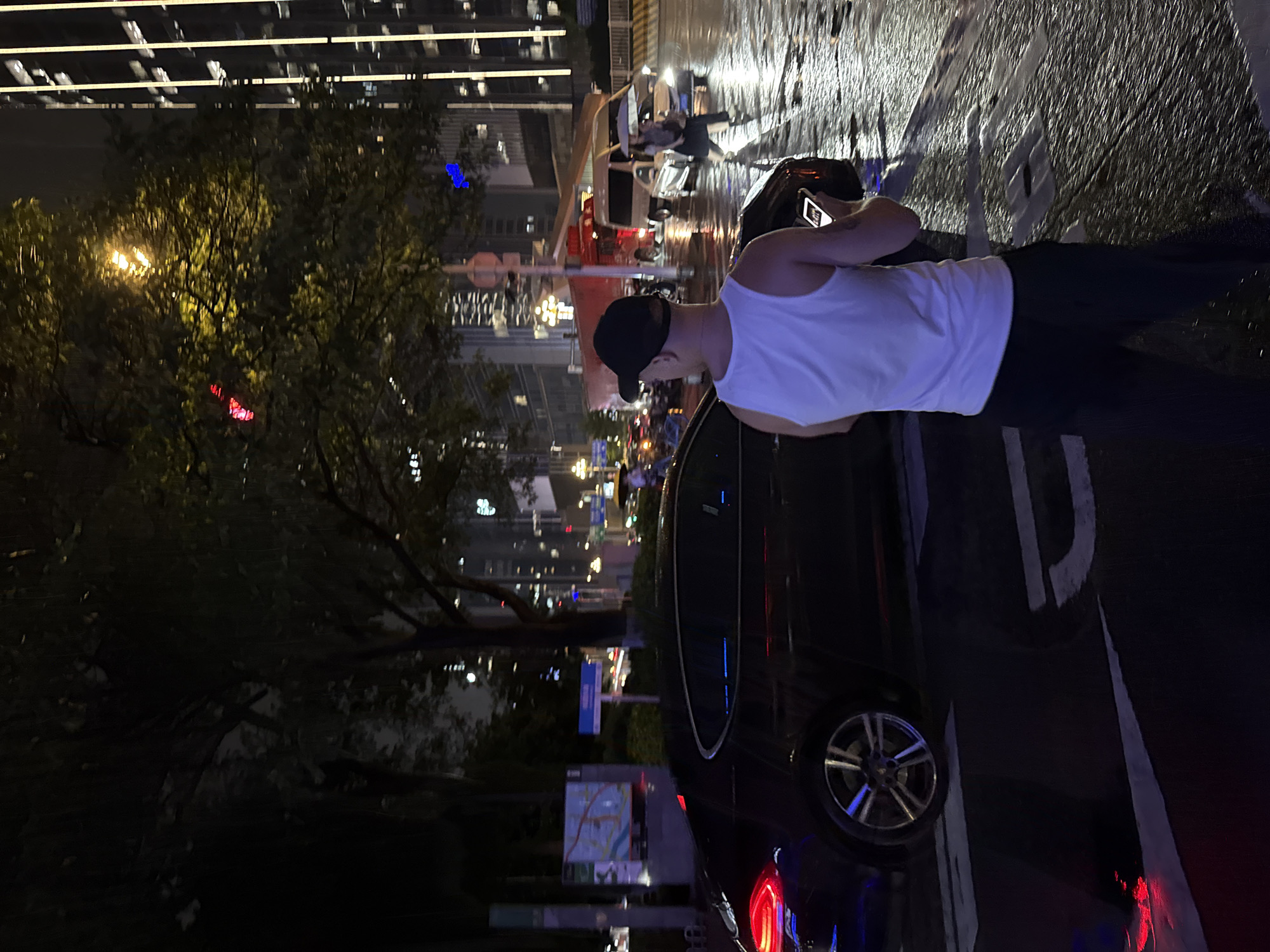 At night in Tianhe District, China, a person wearing a white tank top and a black baseball cap stands on a wet street, looking at a smartphone. The street reflects the lights of the city, including the illuminated windows of tall buildings in the background. A dark sedan is parked to the left, its taillights glowing red. To the right, a crosswalk is visible, and several vehicles, including a van and a red truck, are on the street. Two individuals with an umbrella are crossing the street in the distance. The scene is illuminated by streetlights and the lights from the surrounding buildings, creating a somewhat hazy and reflective atmosphere due to the wet conditions.