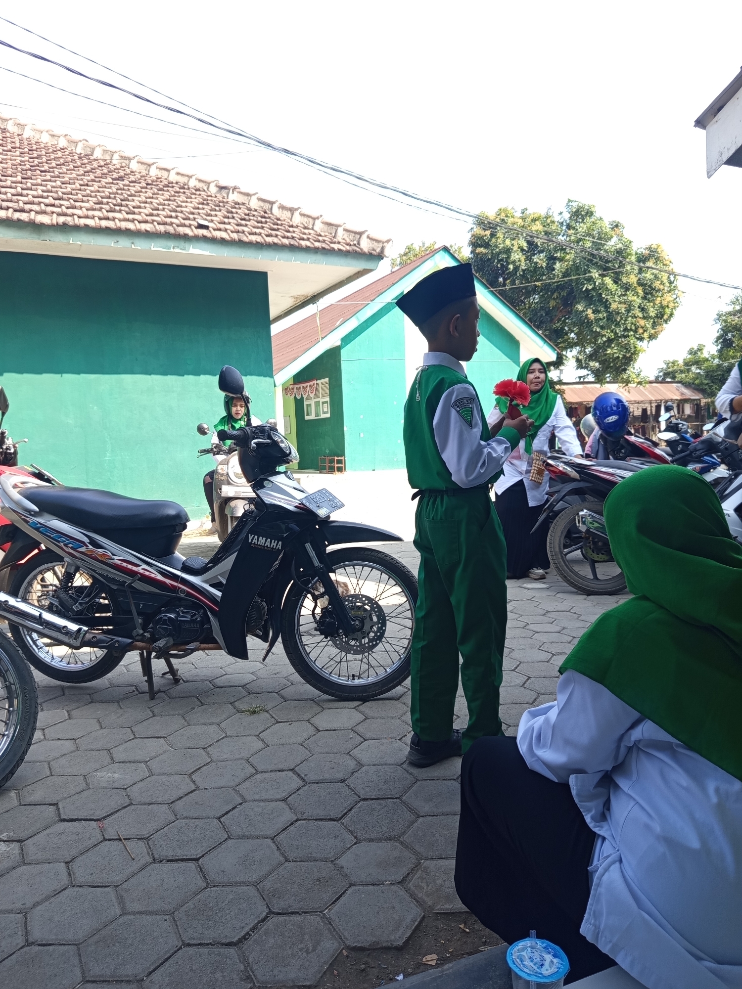 In Lumutan, Indonesia, a young boy in a green and white uniform and a black cap stands facing left, holding red flowers. He is on a paved area in front of a green building. A Yamaha motorcycle is parked to his left, and another motorcycle is visible behind him with a person in a green hijab. In the foreground, a person in a green hijab and white clothing is seated, facing away from the camera. The environment suggests an outdoor setting, possibly a schoolyard or community area, with parked motorcycles indicating transportation.