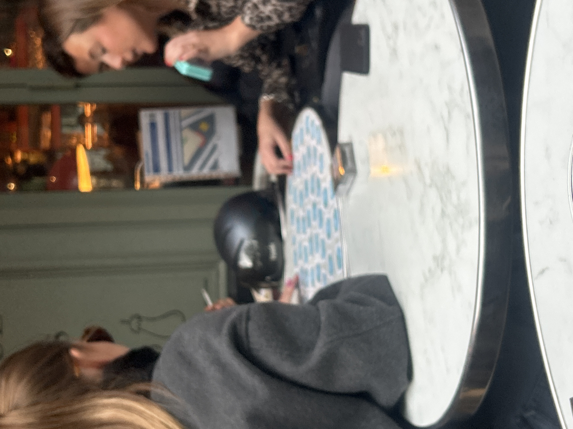 A medium shot captures two women seated at separate round tables in a restaurant or cafe, though the specific location within the city of Unknown, Unknown is unspecified. The woman on the upper left has brown hair and is wearing a leopard-print top, looking at a blue phone or similar device in her hand. Her other hand is resting on a white and blue patterned object, possibly a menu. The woman on the lower left has blonde hair and is wearing a grey sweater. A black object, possibly a purse or helmet, is placed between them on the table. The tables are round with a white marble-like surface and a metallic rim. The backdrop is a blurry mix of interior elements including lighting and what appears to be a sign.