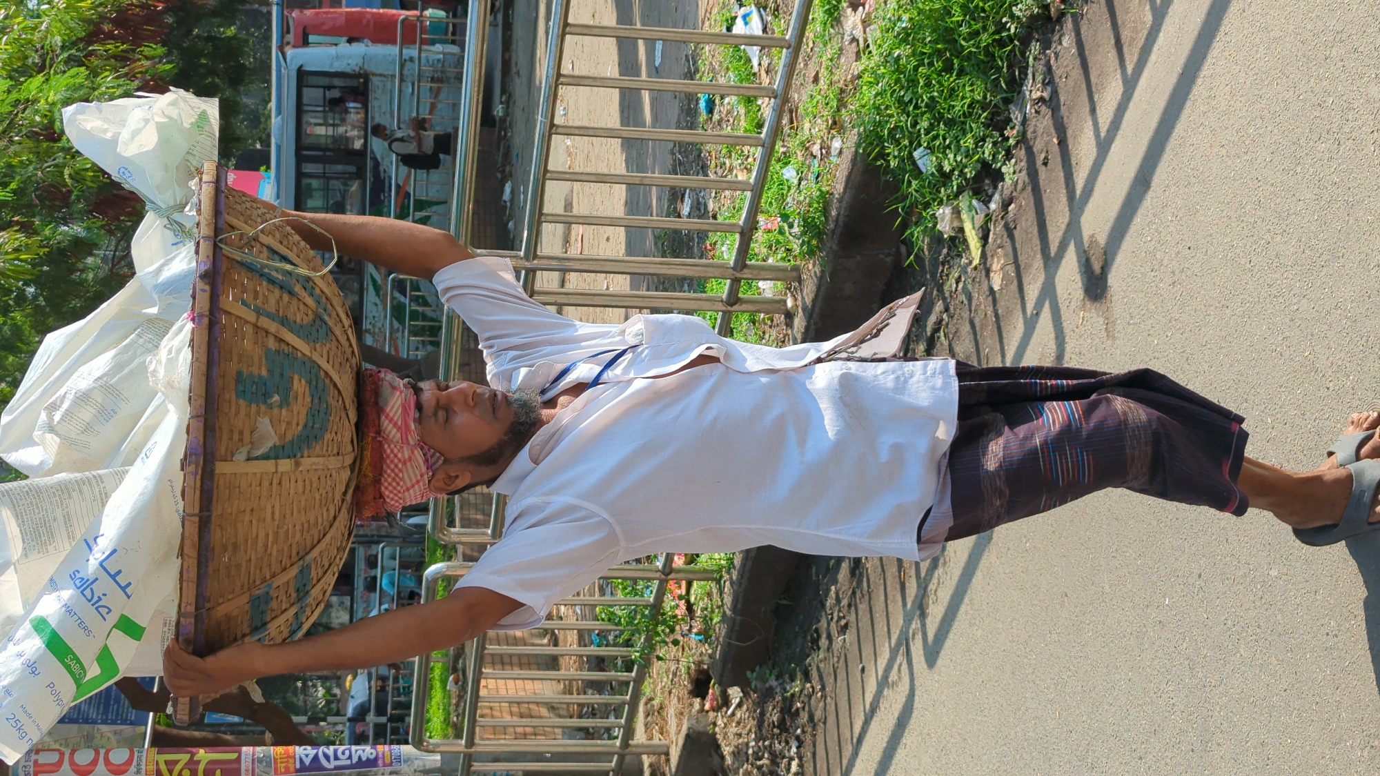 A man in Dhaka, Bangladesh carries a large, woven basket laden with white sacks on his head. The basket's rim is blue and purple. He wears a white shirt, a dark-colored lungi (traditional garment), and gray sandals. A red and white patterned cloth is wrapped around his head beneath the basket. He walks on a paved surface next to a metal railing with green foliage beyond it. A bus with people inside is visible in the background. A sign with Bengali script is also visible near the bottom of the frame. The sacks on the basket are marked with "SABIC" logo and Bengali script.