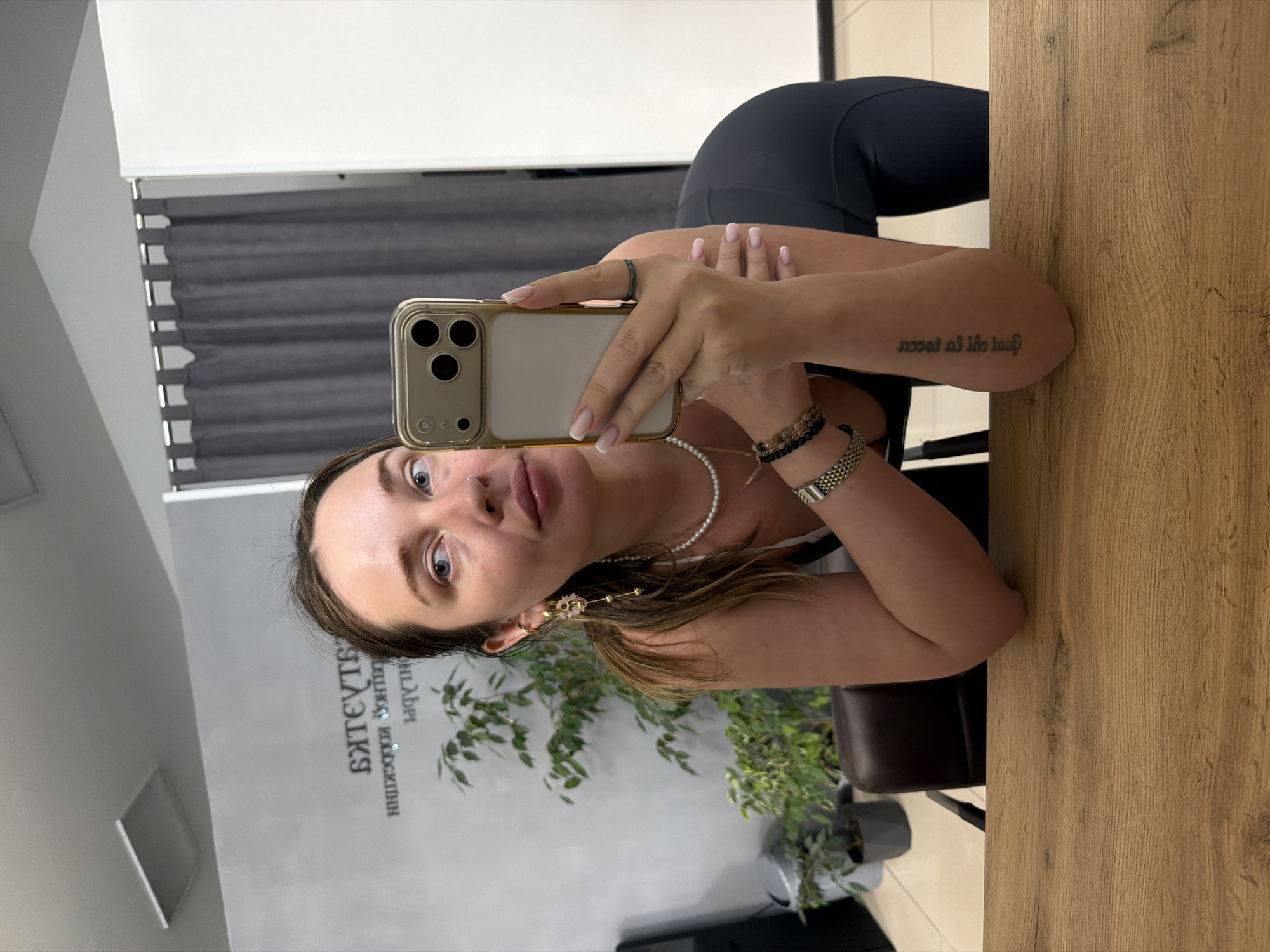 A female subject is depicted in a mirror selfie taken indoors within a commercial establishment in Chaikovsky, Russia. She is leaning forward, resting her elbows on a light brown wooden table, while holding a gold-framed smartphone with three prominent camera lenses in her right hand to capture her reflection. The subject has light brown hair, blue eyes, and wears a dark top over a white layer, and dark leggings. She is adorned with a pearl necklace, a gold chain, multiple bracelets on her left wrist, gold-colored dangle earrings with a floral design, and a dark green ring on her right ring finger. A small stud nose piercing is visible on her left nostril, and her fingernails display a French tip manicure. Text is tattooed on her right forearm. The background, reflected in the mirror, features a light gray wall with reversed Russian text, "БЬЮТИ КОНФЕКШН..." (Beauty Confection/Collection), suggesting a salon or boutique. A green potted plant, a dark brown office chair, a light-colored floor, and a section with dark gray curtains are also visible.