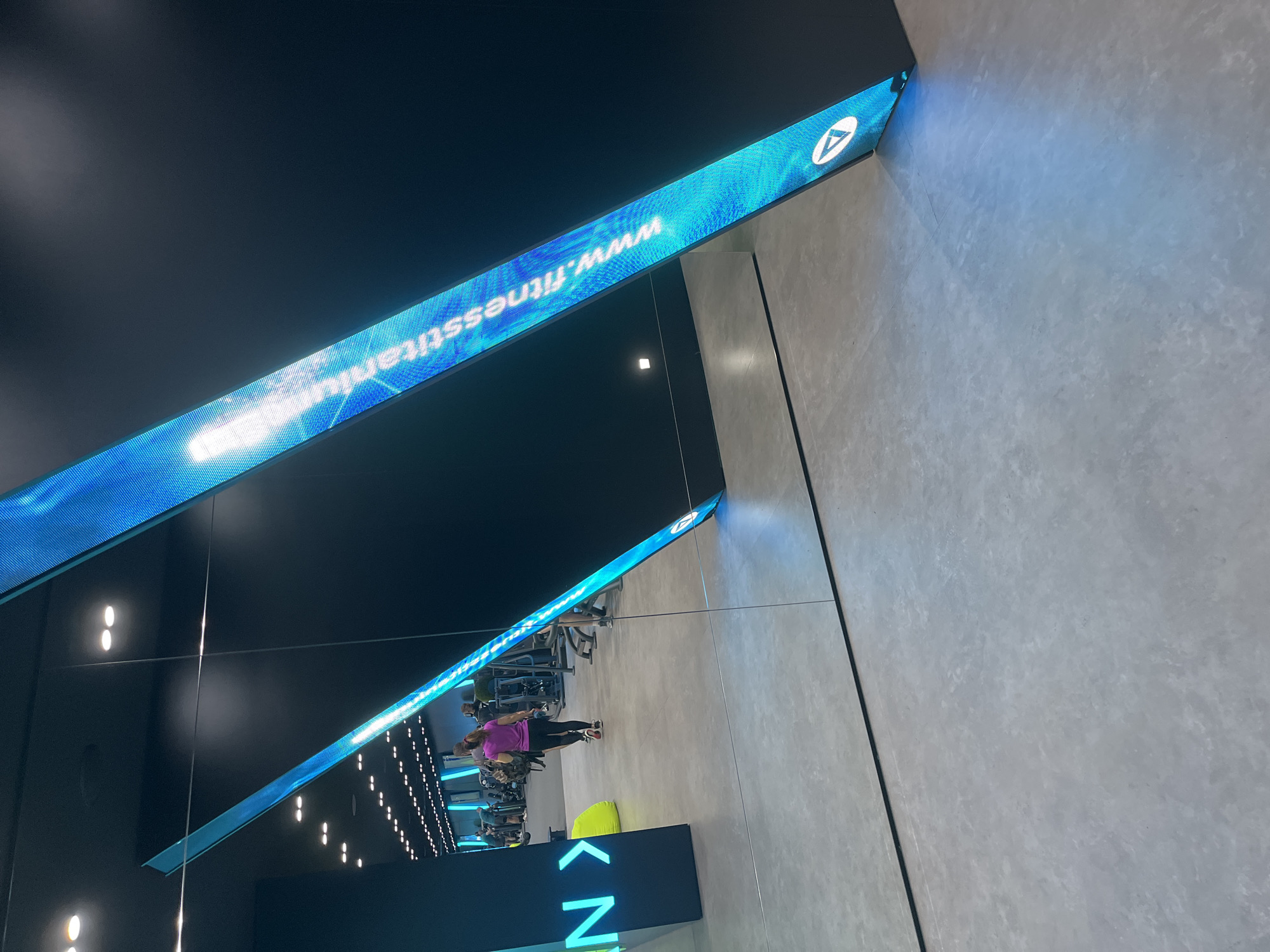 Here is a description of the image:
The image appears to be taken inside a gym, likely in the city of Unknown. A long, blue LED strip with the website "www.fitnesstitanium.com" runs along the top of a black wall, mirroring the reflection of another LED strip below.
The reflection shows people exercising on equipment. The interior is brightly lit with rows of lights along the ceiling, suggesting a modern fitness center. A sign with a large arrow and the letter "N" is visible, possibly indicating the direction of certain areas within the gym. The floor is tiled with light gray material.