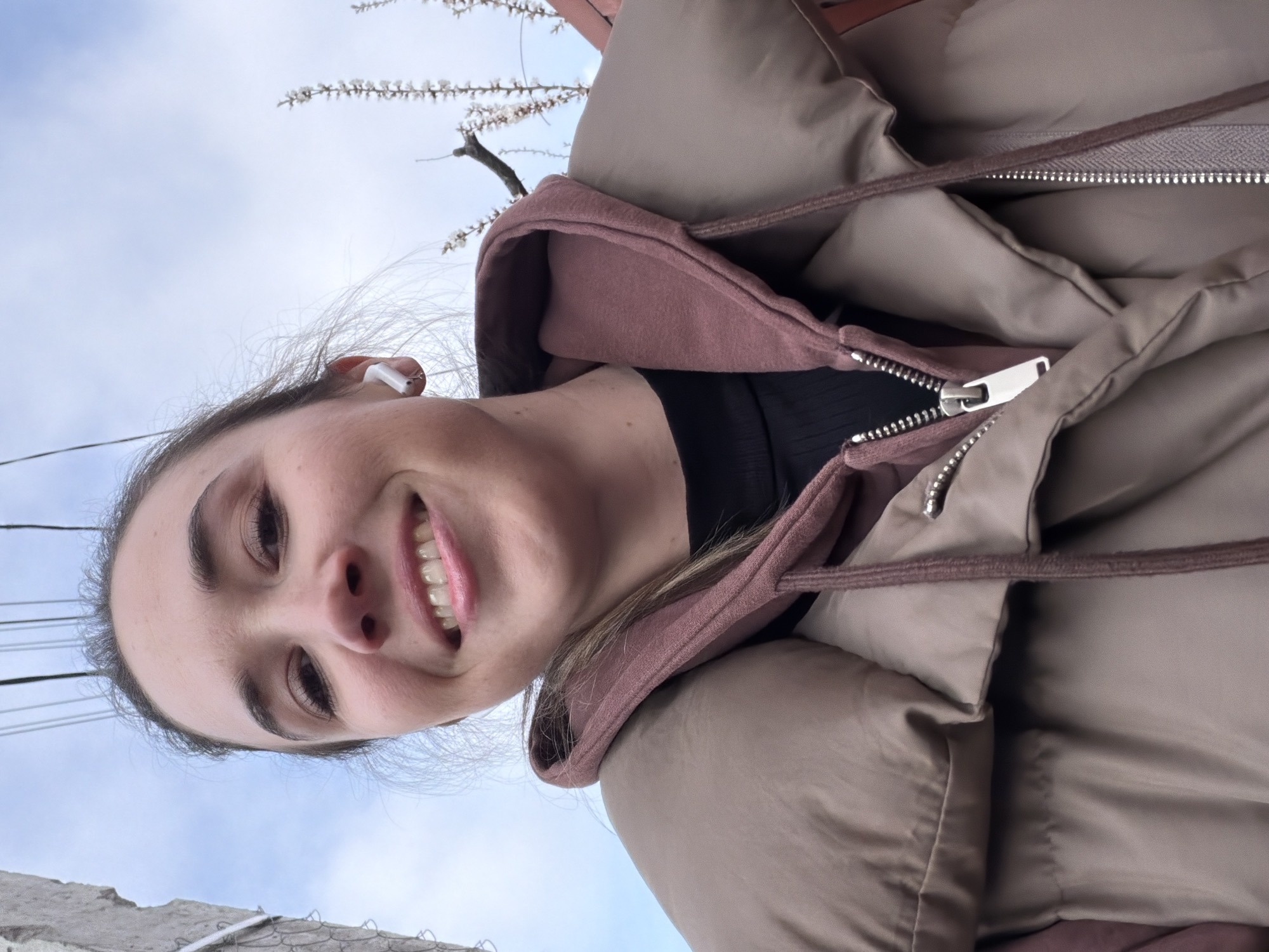 The image is an upward-angled selfie of a smiling young woman with fair skin and brown hair. She is wearing a pink hoodie under a beige puffer jacket. She has a white wireless earbud in her left ear. The background shows a pale blue sky, the top of a concrete structure, and a blossoming tree branch. Considering the request to locate the scene in Rostov-on-Don, Russia, the concrete structure could be a part of a building or infrastructure common in urban areas, and the blooming branch suggests it might be springtime. The low angle and close-up focus on the woman obscure the possibility of definitively identifying a specific location. The setting seems to be outdoors, possibly in a park or urban area with trees and buildings. The general style and content suggest the image was captured recently.