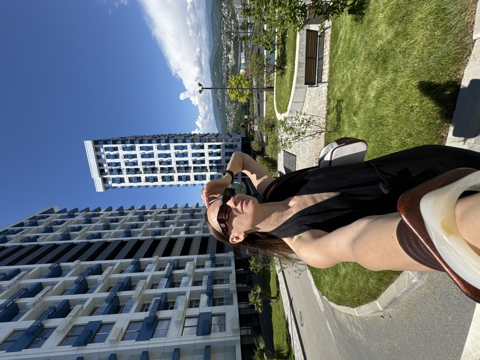 A selfie-style image captures a woman in a black top and rectangular sunglasses, shielding her eyes with her right hand, holding a phone or camera in her left hand, with a partial view of her left arm and a white and brown object in the immediate foreground. She is positioned outdoors on a sunny day.
The background features two modern, tall residential buildings with white facades and prominent blue balcony details. The building on the left has visible air conditioning units. A well-maintained urban green space is visible, including a curved grassy median, small trees, bushes, and a paved pathway with a wooden bench. A street light with two luminaires stands in the mid-ground. In the far background, a sprawling cityscape extends across undulating hills, culminating in a range of distant mountains under a clear blue sky with some scattered cumulus clouds. The presence of palm trees near the base of the left building, the modern high-rise architecture, and the mountainous backdrop confirm the location as the city of Sochi, Russia.