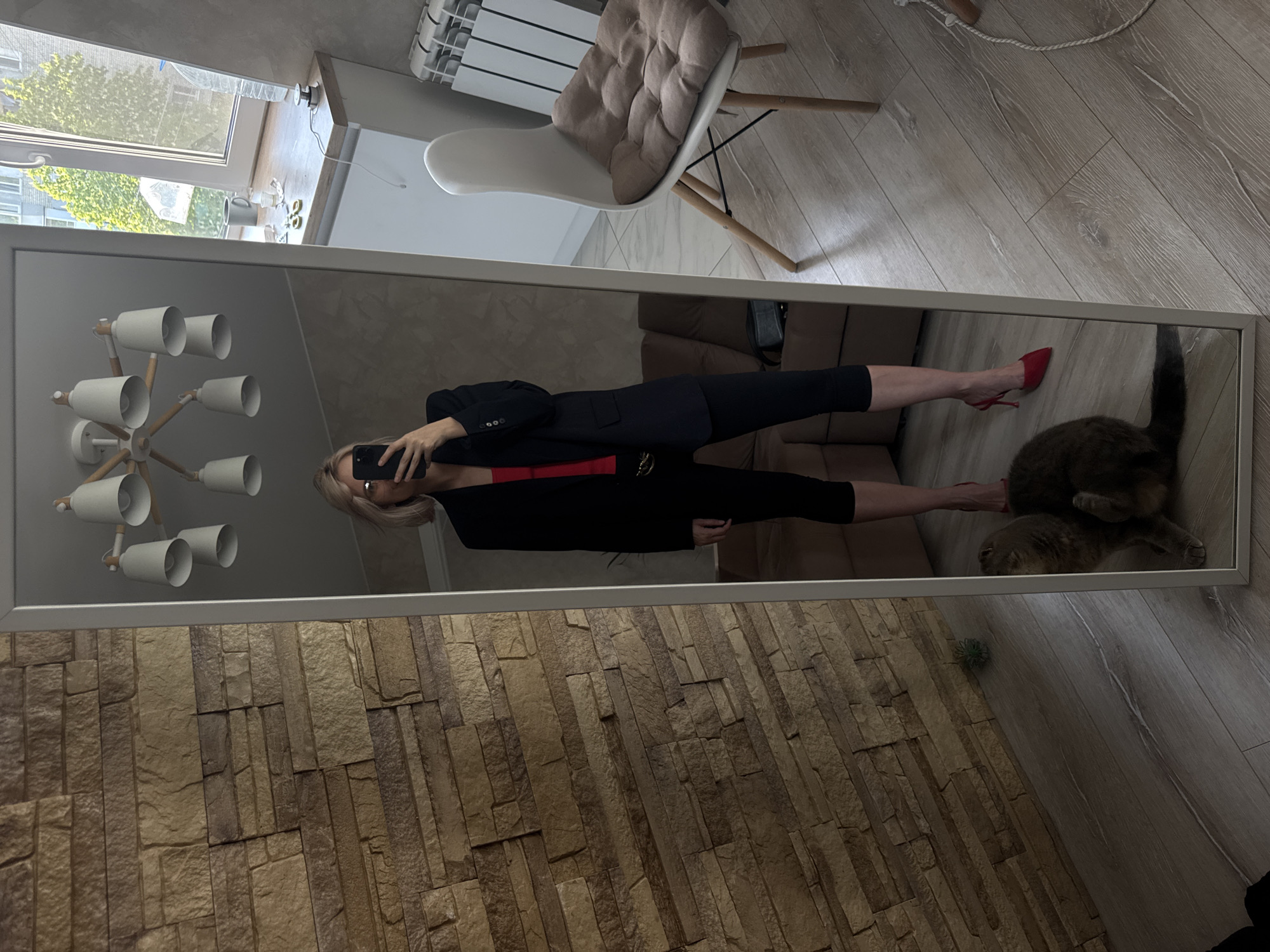 A woman in Saratov, Russia takes a mirror selfie in an indoor location with a cat present. The woman is dressed in black leggings, a red top, and a navy blazer. She is holding her phone to take the picture, partially obscuring her face. The mirror reflects a modern chandelier on the ceiling, and a living room setting with a sofa. Near the bottom of the mirror, a brown cat is visible, close to the floor. The environment also includes a wall with brick-like pattern, wooden flooring and a chair is partially visible in the upper part of the image. Outside the window, other buildings and a tree can be seen.